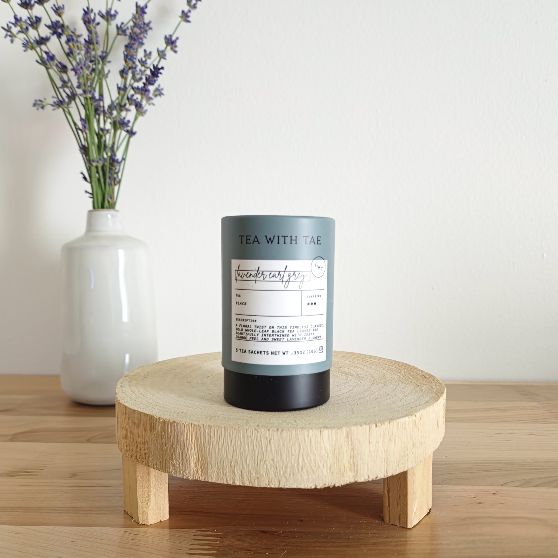 A cylindrical tea canister labeled "Tea With Tae, Lavender Earl Grey Black Tea" stands on a light wooden display stand. In the background, a white vase holds a small bouquet of lavender sprigs, against a plain white wall.