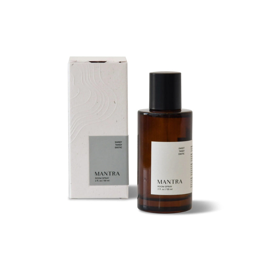 Brown glass bottle of Mantra Room Spray, with black spray nozzle and cap, resting on a light-colored textured surface. The bottle's label reads 'Mantra'. A small gold dish holding a dark stone and a white crystal is positioned to the right of the bottle. A portion of a round mirror and a light-colored wall are visible in the background.