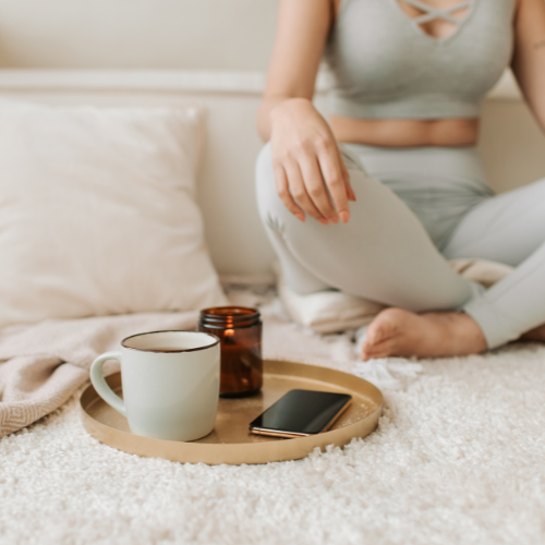 A woman sits cross-legged on a bed, wrapped in light-colored blankets, holding a mug of tea with both hands. She is wearing a grey ribbed top and leggings, and a gold tray with candles is visible beside her. The scene evokes a sense of peaceful self-care.