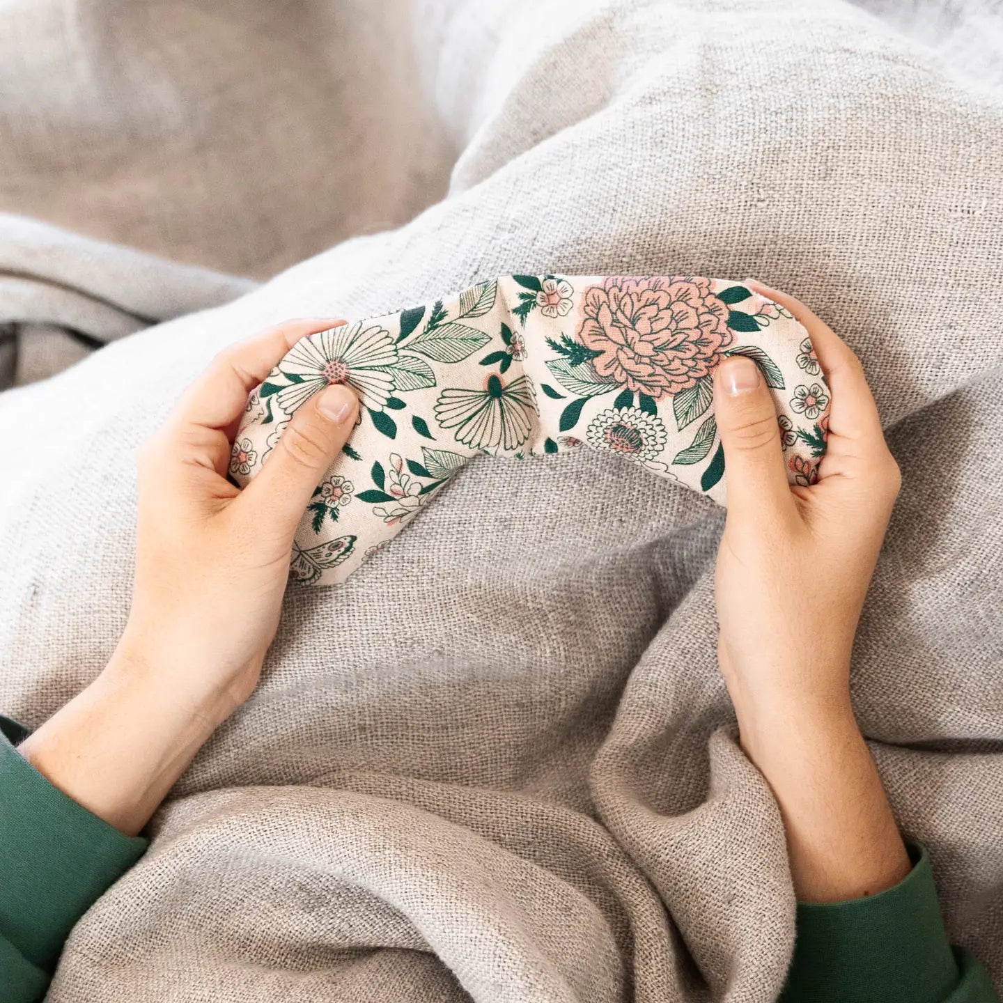 Hands gently hold a floral-patterned eye pillow with a design of pink peonies and green leaves, resting on light grey linen bedding. The image suggests comfort and relaxation.