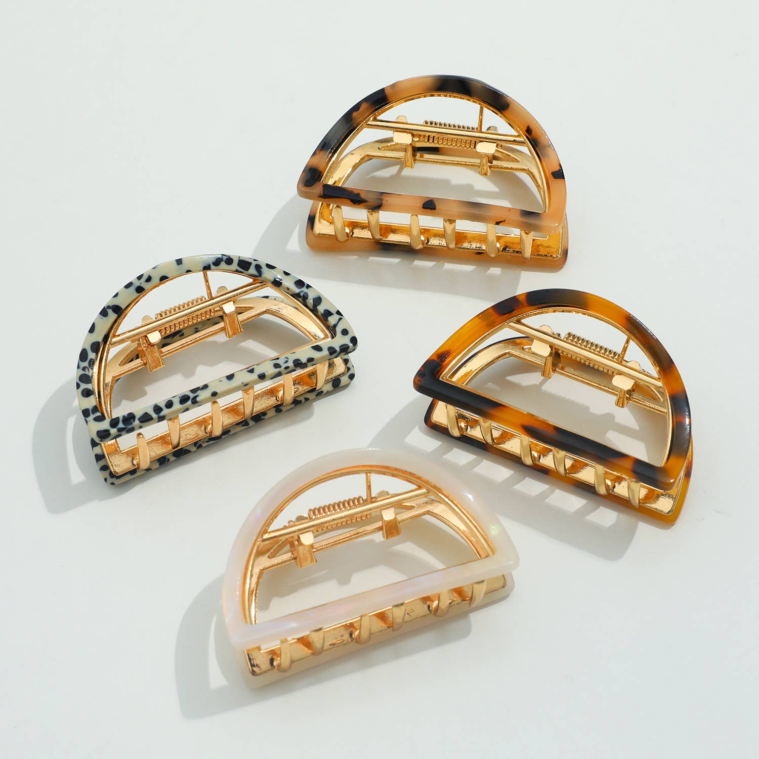 Four D-shaped hair claw clips are arranged on a white surface, featuring various patterns: classic tortoise shell, a speckled Dalmatian print, a lighter translucent tortoise shell, and a creamy white iridescent finish. All clips have gold-toned hardware.