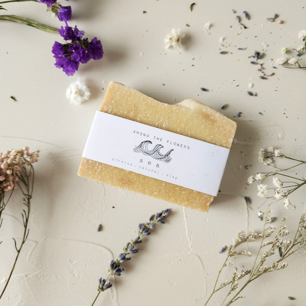 A rectangular bar of light yellow "Sea" natural soap, with a label featuring a wave design, rests on a light textured surface. It is surrounded by scattered dried flowers in shades of purple, white, and muted pink, creating a natural, botanical aesthetic.