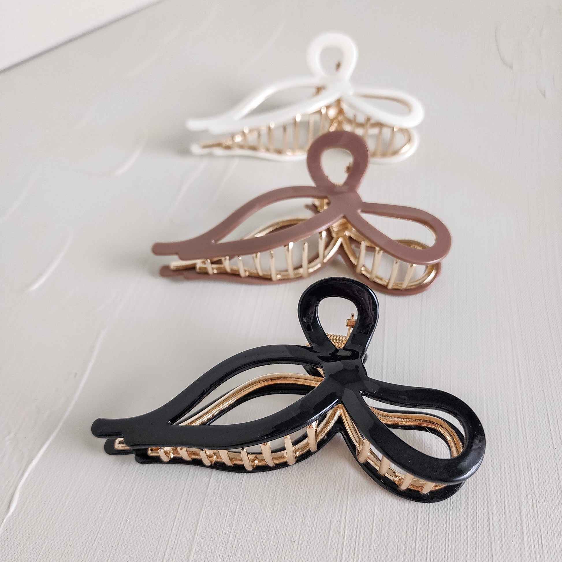 Three bow-shaped metal claw clips in black, brown, and white are neatly aligned on a light textured surface. The clips feature gold-toned inner clasps, showcasing their elegant design.