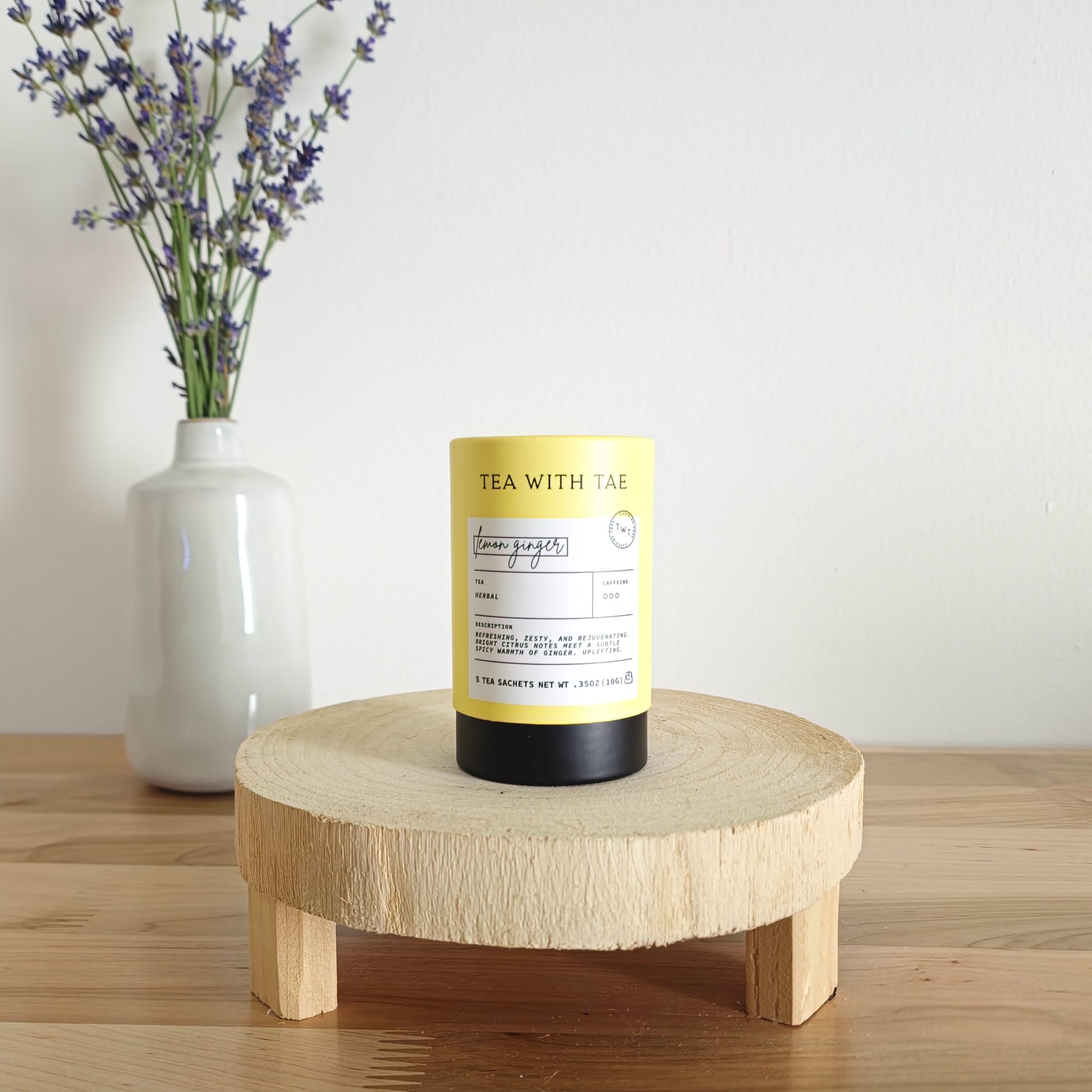 A cylindrical tea canister labeled "Tea With Tae, Lemon Ginger Herbal Tea" stands on a light wooden display stand. In the background, a white vase holds a small bouquet of lavender sprigs, against a plain white wall.