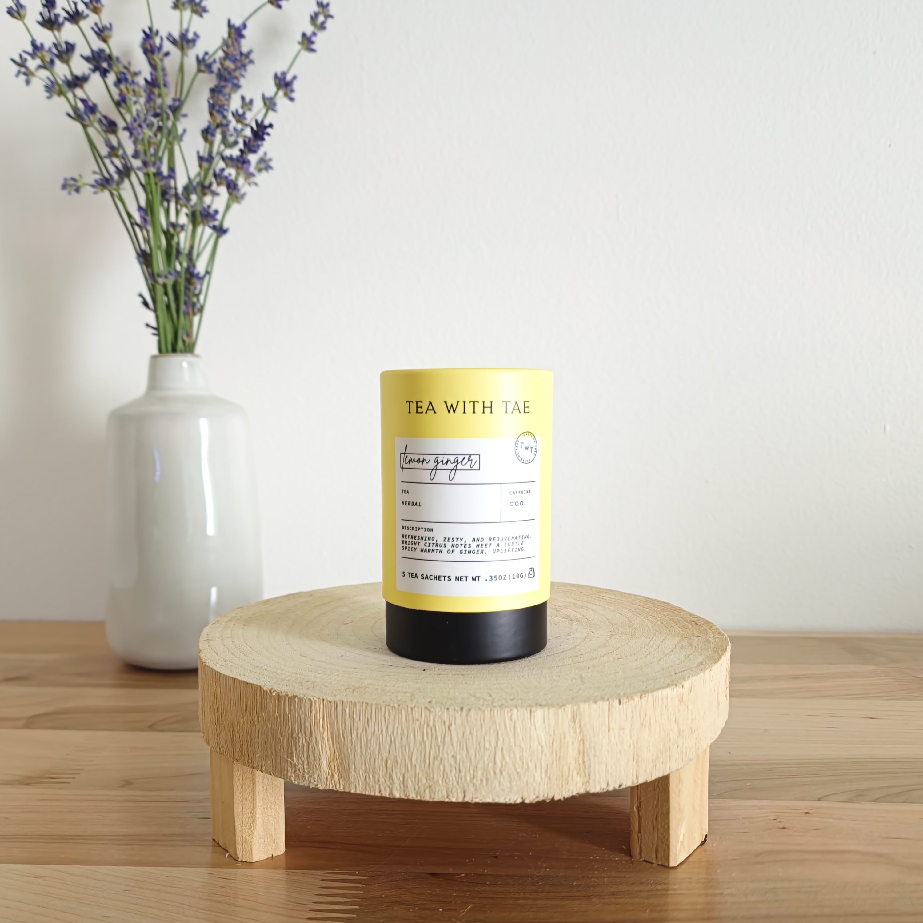 A cylindrical tea canister labeled "Tea With Tae, Lemon Ginger Herbal Tea" stands on a light wooden display stand. In the background, a white vase holds a small bouquet of lavender sprigs, against a plain white wall.