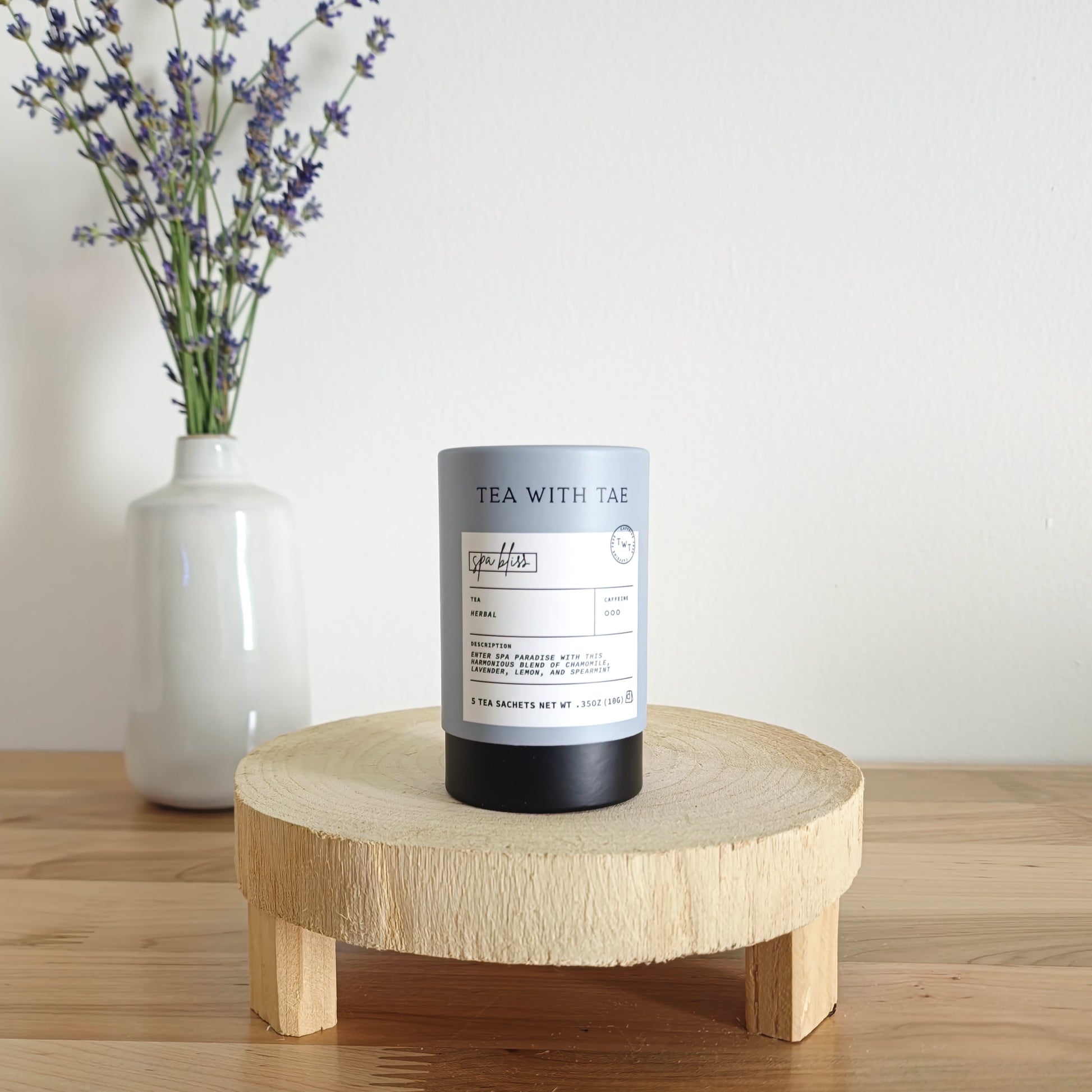 A cylindrical tea canister labeled "Tea With Tae, Spa Bliss Herbal Tea" stands on a light wooden display stand. In the background, a white vase holds a small bouquet of lavender sprigs, against a plain white wall.