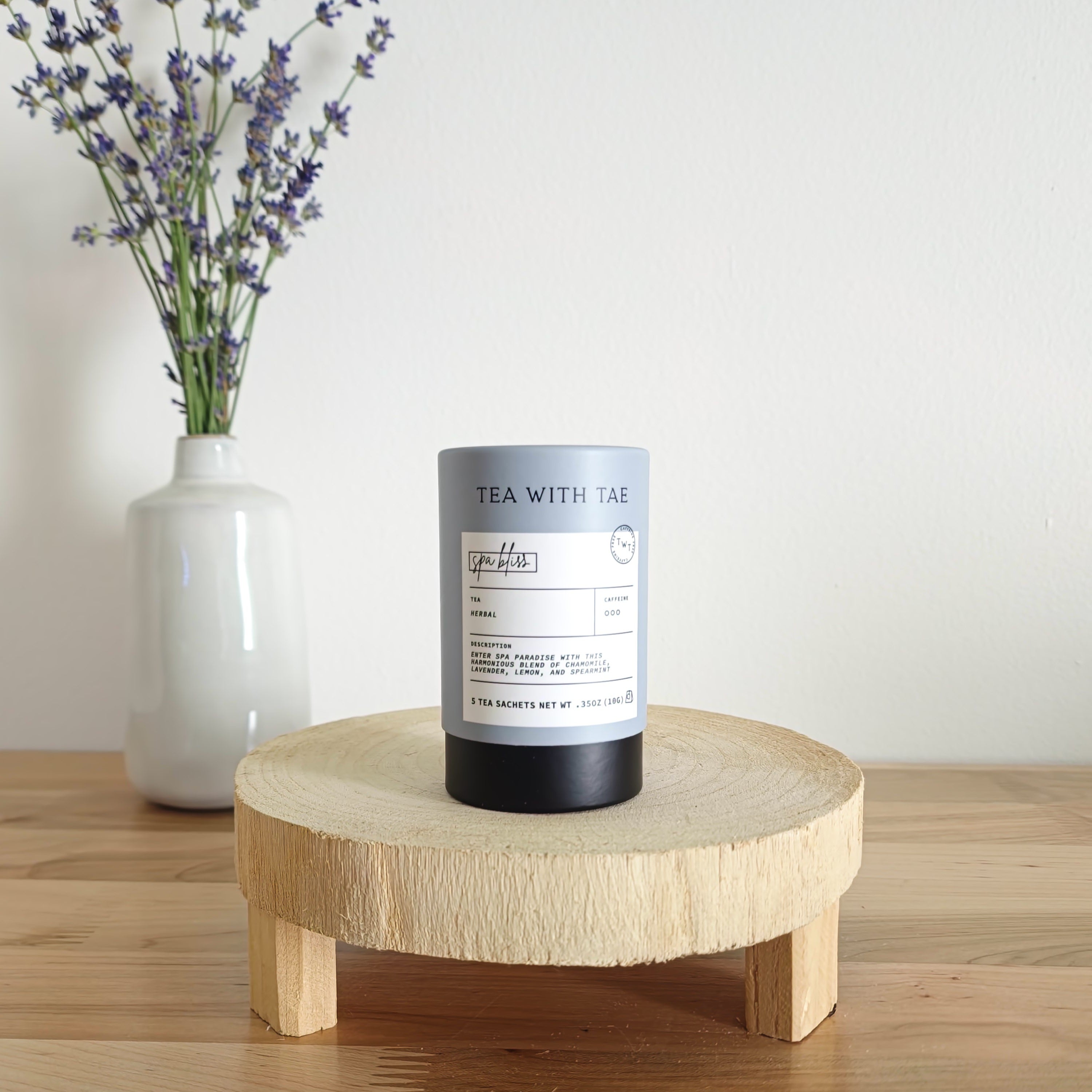 A cylindrical tea canister labeled "Tea With Tae, Spa Bliss Herbal Tea" stands on a light wooden display stand. In the background, a white vase holds a small bouquet of lavender sprigs, against a plain white wall.