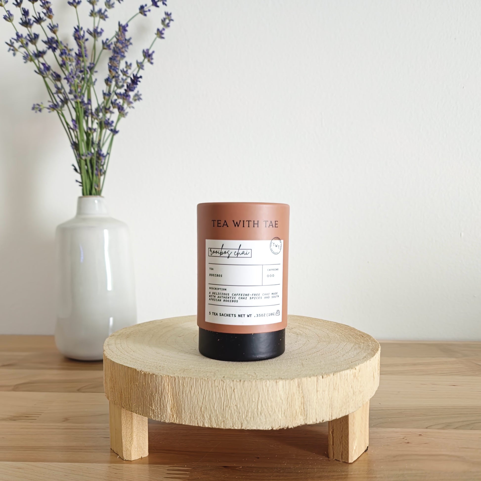 A cylindrical tea canister labeled "Tea With Tae, Bombay Chai Rooibos" stands on a light wooden display stand. In the background, a white vase holds a small bouquet of lavender sprigs, against a plain white wall.
