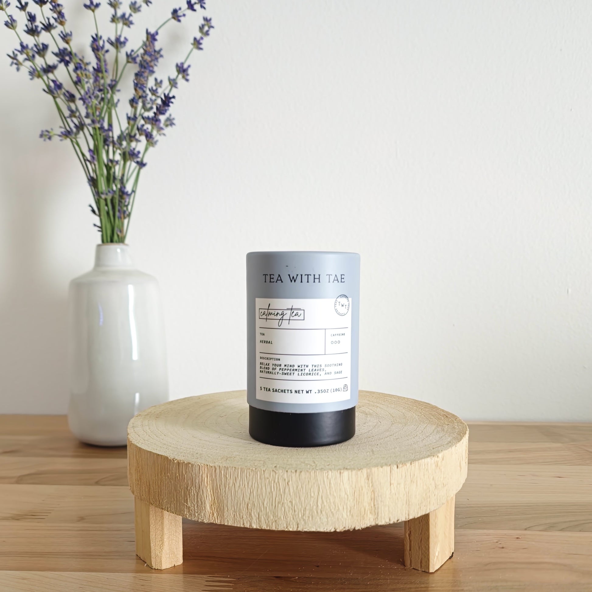  A cylindrical tea canister labeled "Tea With Tae, Calming Tea Herbal" stands on a light wooden display stand. In the background, a white vase holds a small bouquet of lavender sprigs, against a plain white wall.