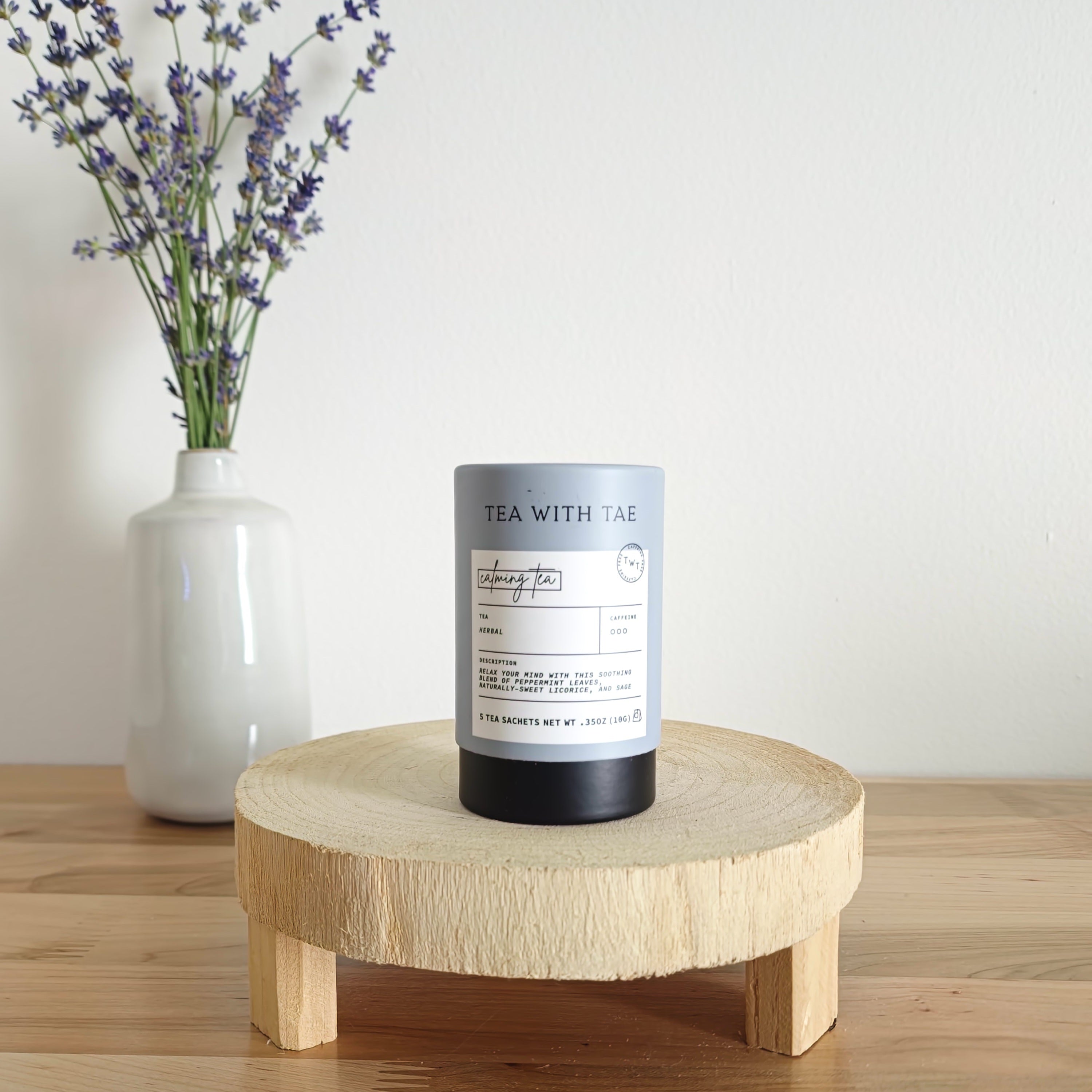  A cylindrical tea canister labeled "Tea With Tae, Calming Tea Herbal" stands on a light wooden display stand. In the background, a white vase holds a small bouquet of lavender sprigs, against a plain white wall.