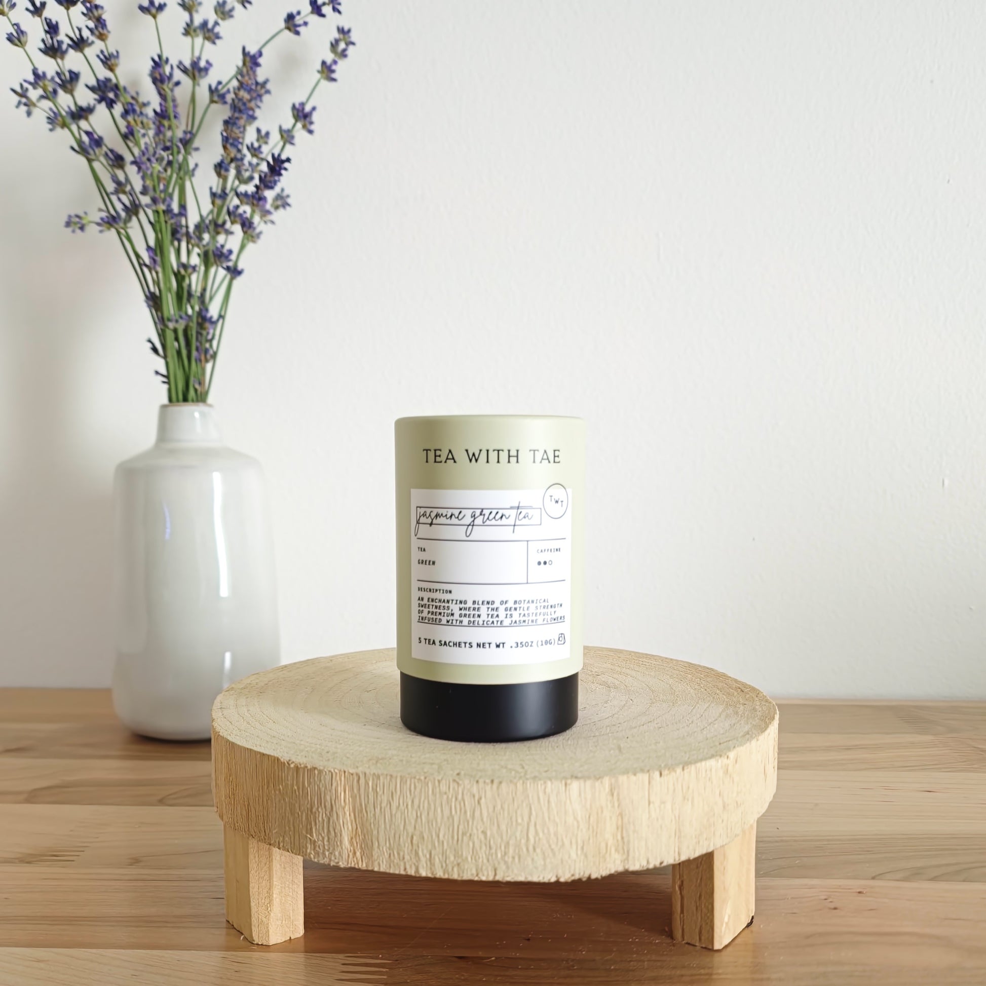 A cylindrical tea canister labeled "Tea With Tae, Jasmine Green Tea" stands on a light wooden display stand. In the background, a white vase holds a small bouquet of lavender sprigs, against a plain white wall.