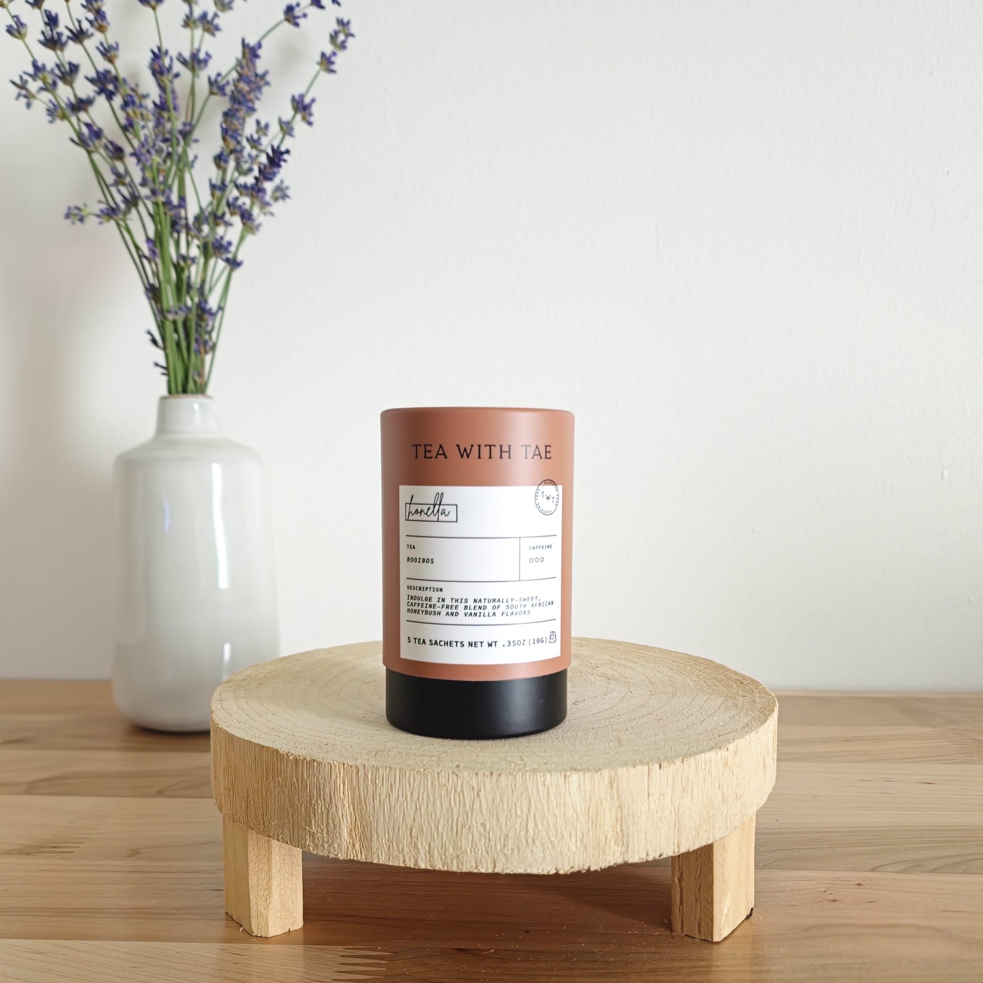 A cylindrical tea canister labeled "Tea With Tae, Rooibos" stands on a light wooden display stand. In the background, a white vase holds a small bouquet of lavender sprigs, against a plain white wall.