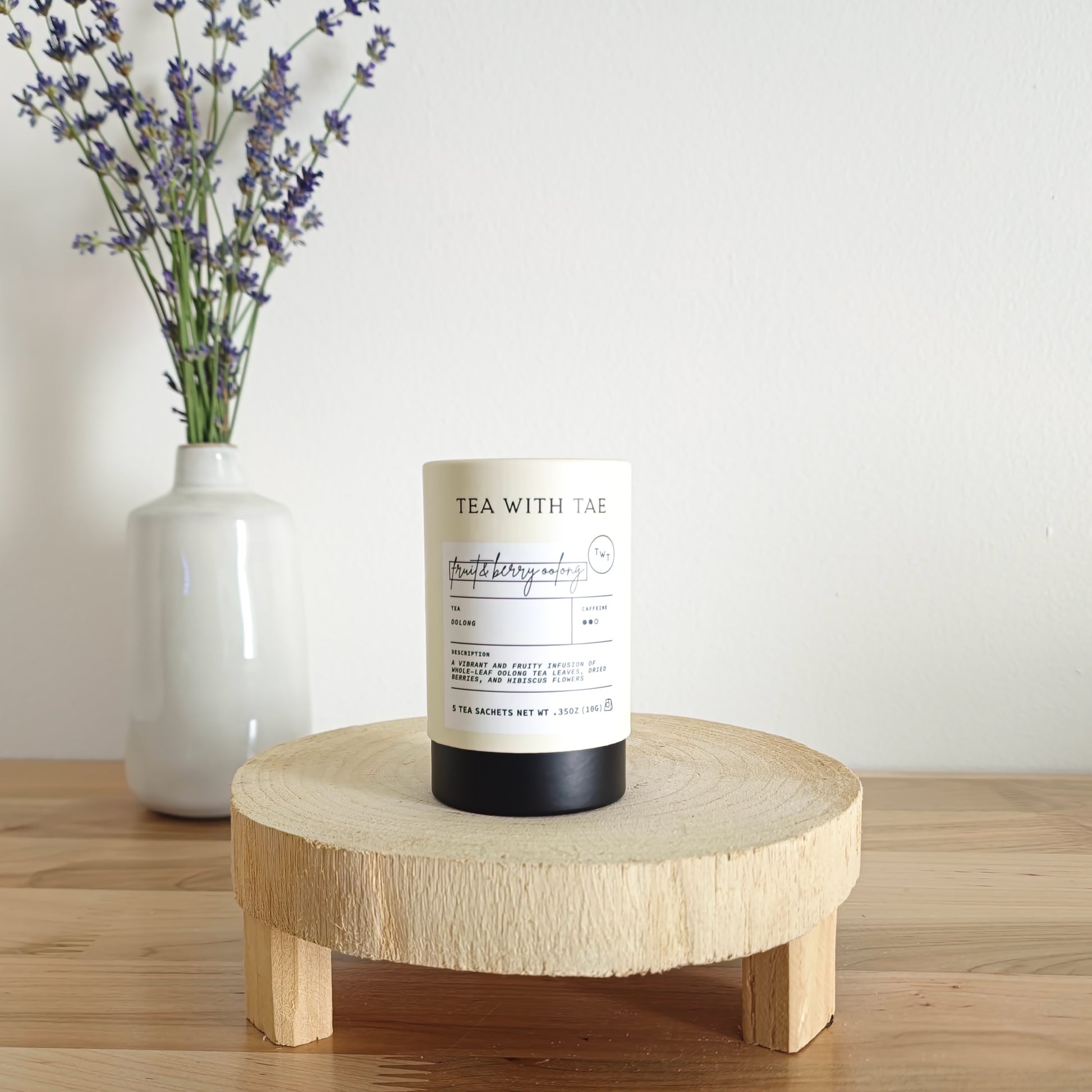 A cylindrical tea canister labeled "Tea With Tae, Wild Berry Oolong Tea" stands on a light wooden display stand. In the background, a white vase holds a small bouquet of lavender sprigs, against a plain white wall.