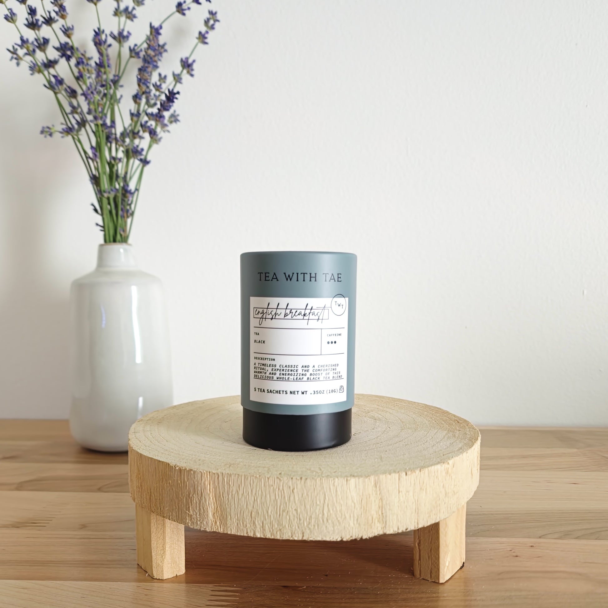 A cylindrical tea canister labeled "Tea With Tae, English Breakfast Black Tea" stands on a light wooden display stand. In the background, a white vase holds a small bouquet of lavender sprigs, against a plain white wall.