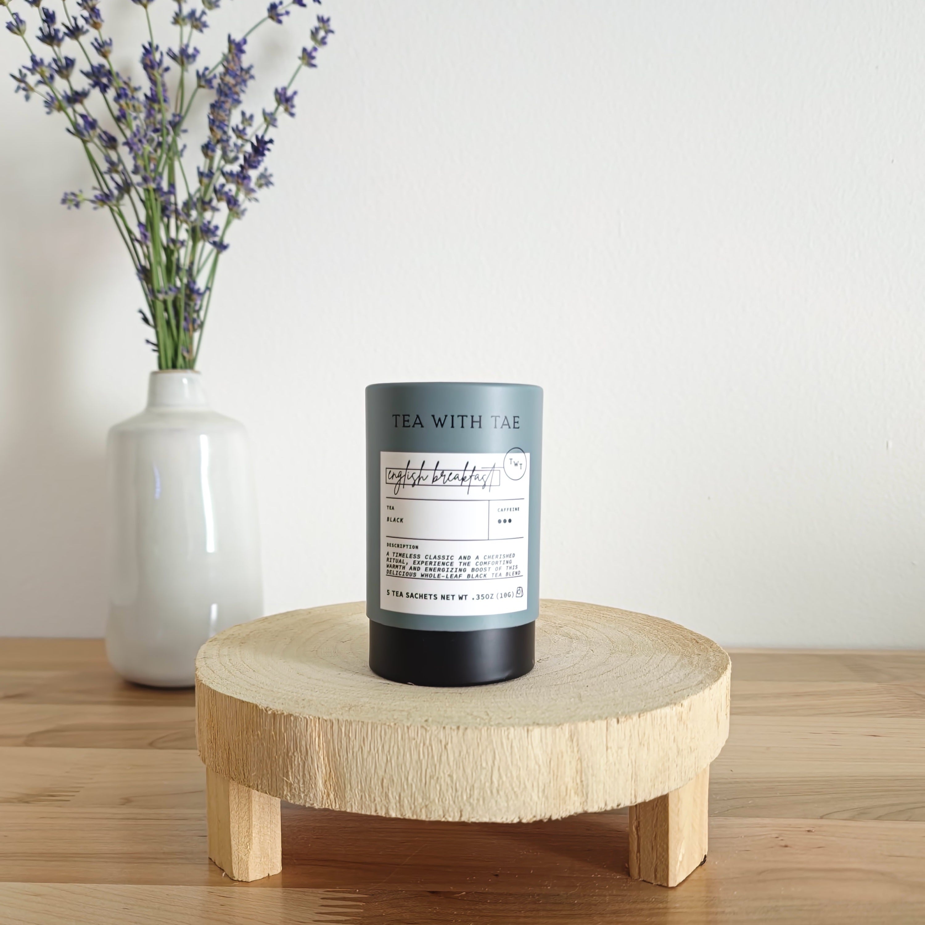 A cylindrical tea canister labeled "Tea With Tae, English Breakfast Black Tea" stands on a light wooden display stand. In the background, a white vase holds a small bouquet of lavender sprigs, against a plain white wall.