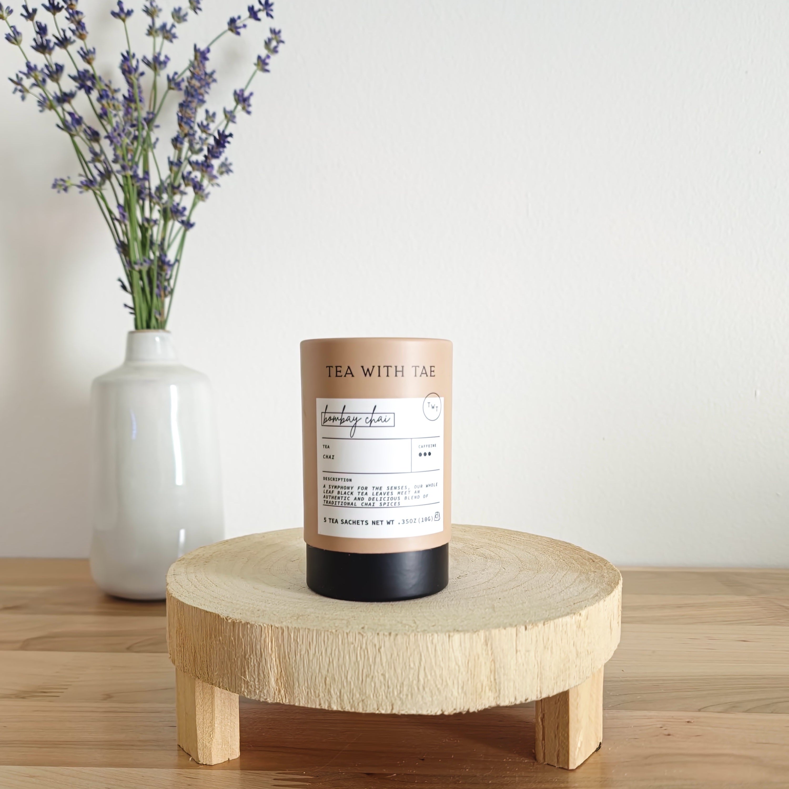 A cylindrical tea canister labeled "Tea With Tae, Bombay Chai Black Tea" stands on a light wooden display stand. In the background, a white vase holds a small bouquet of lavender sprigs, against a plain white wall.