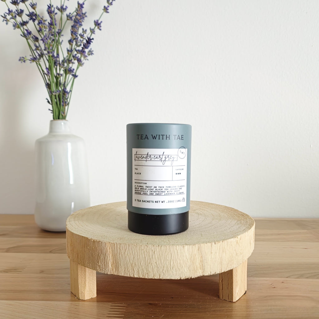 A cylindrical tea canister labeled "Tea With Tae, Lavender Earl Grey Black Tea" stands on a light wooden display stand. In the background, a white vase holds a small bouquet of lavender sprigs, against a plain white wall.