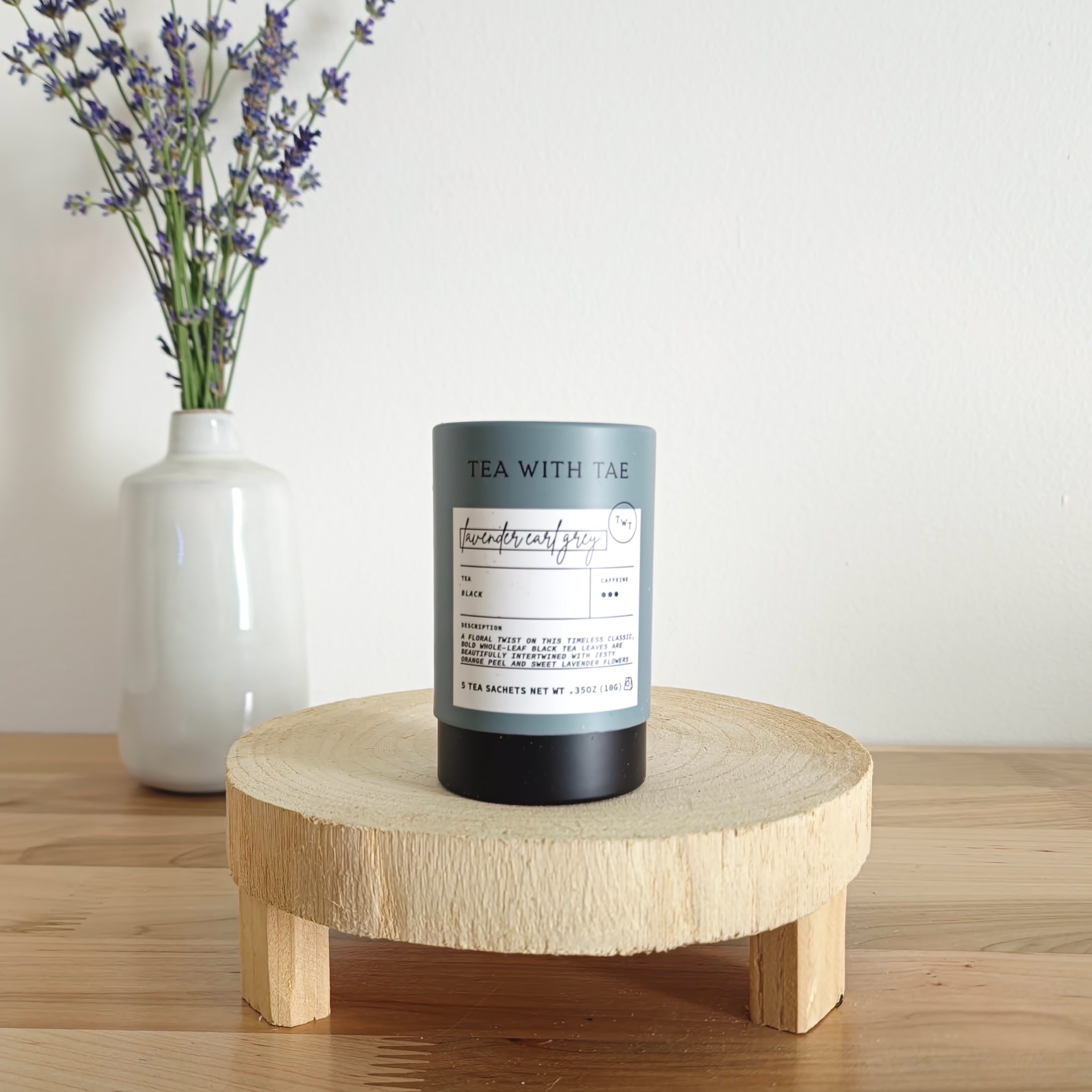 A cylindrical tea canister labeled "Tea With Tae, Lavender Earl Grey Black Tea" stands on a light wooden display stand. In the background, a white vase holds a small bouquet of lavender sprigs, against a plain white wall.