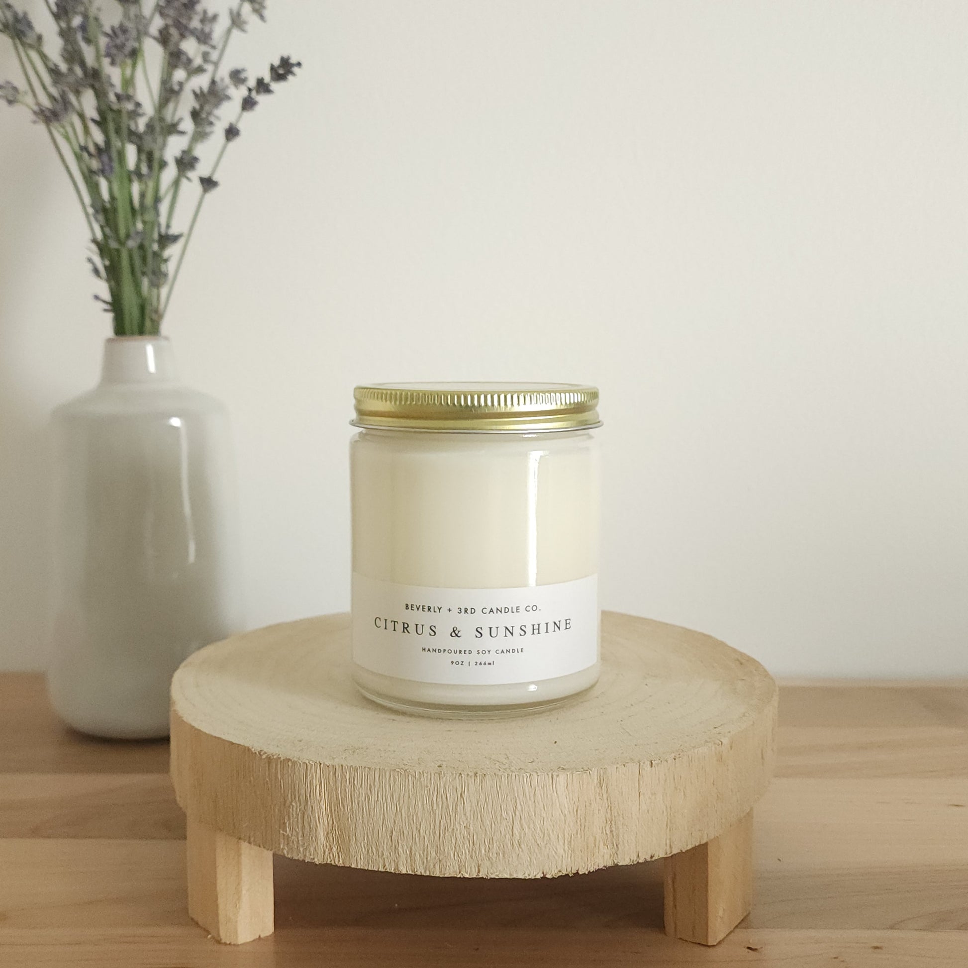 A "Citrus & Sunshine" scented soy candle in a clear glass jar with a gold lid and a white label, sits on a light wooden display stand. In the background, a white vase holds a small bouquet of lavender sprigs, against a plain white wall.