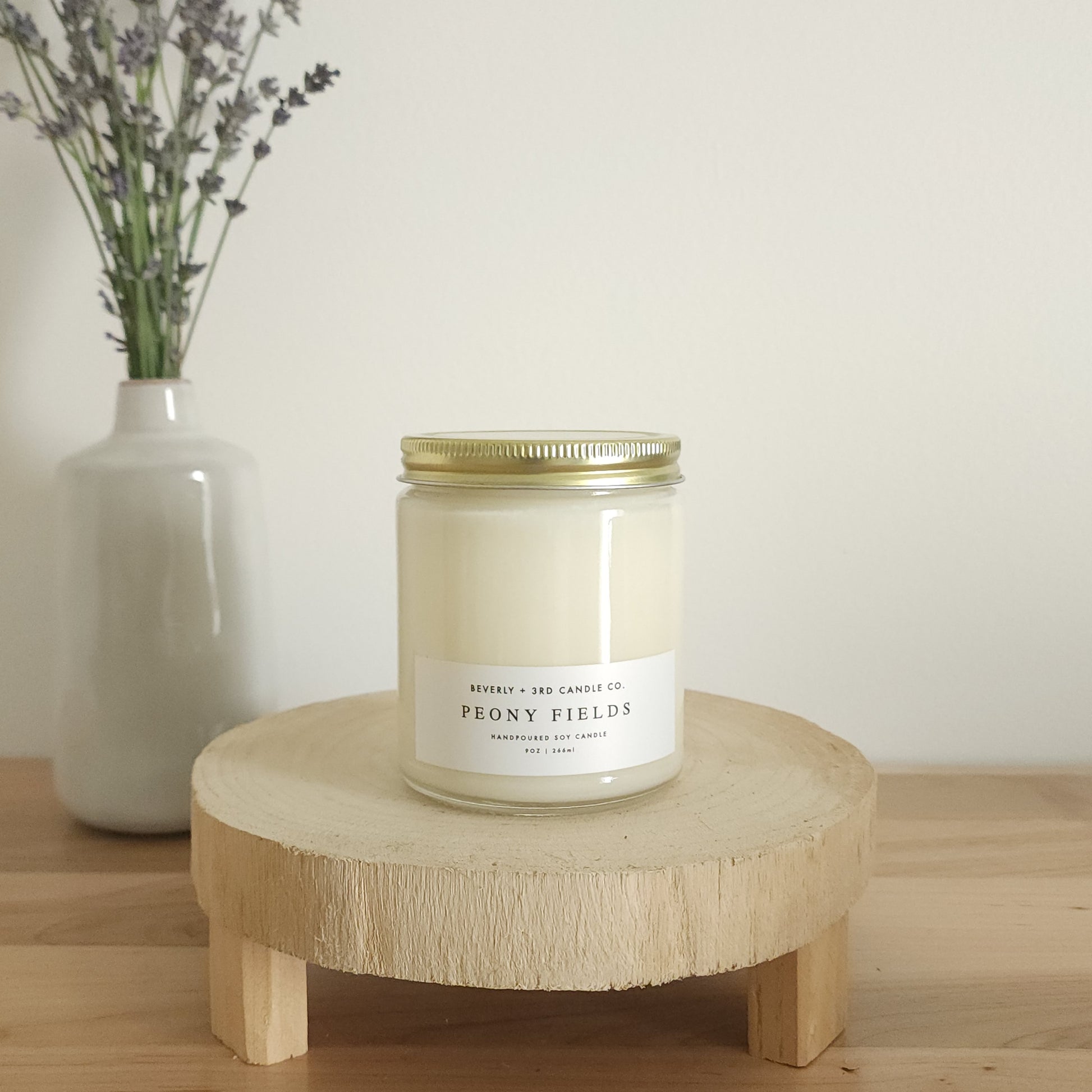 A "Peony Fields" scented soy candle in a clear glass jar with a gold lid and a white label, sits on a light wooden display stand. In the background, a white vase holds a small bouquet of lavender sprigs, against a plain white wall.