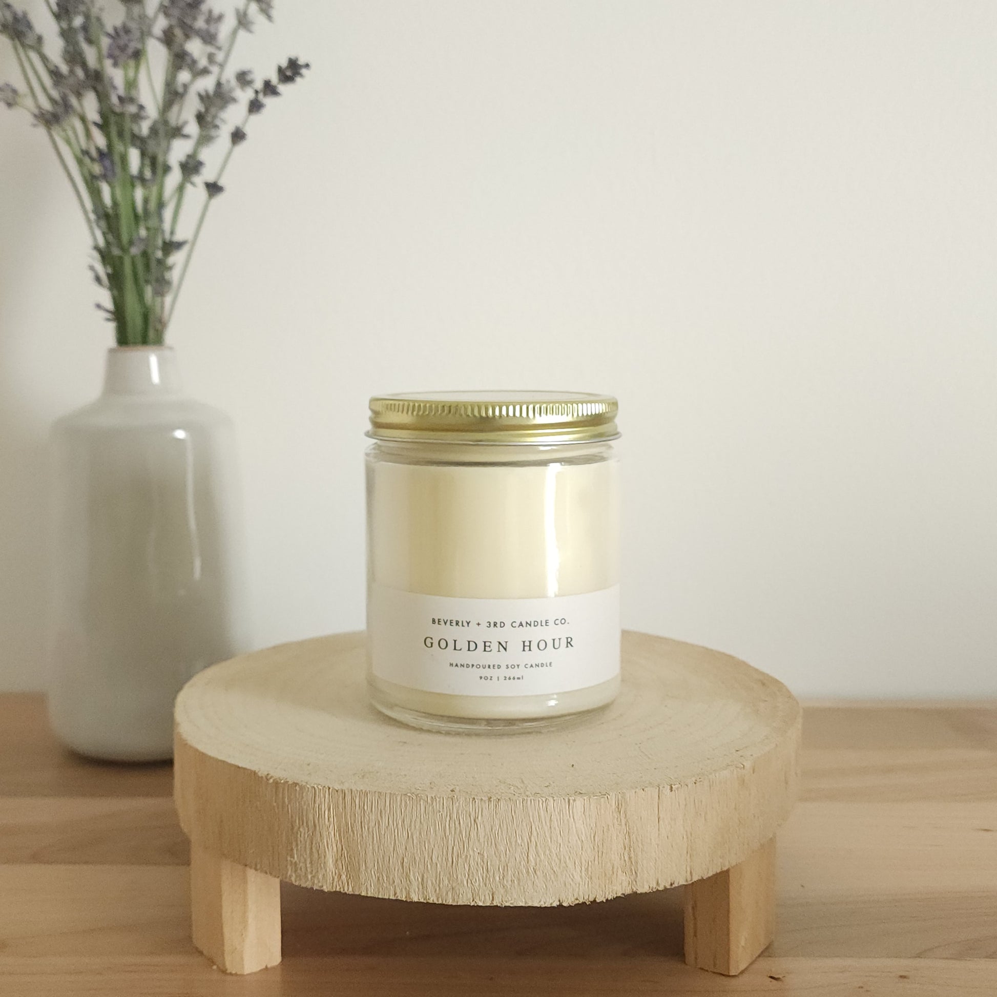 A "Golden Hour" scented soy candle in a clear glass jar with a gold lid and a white label, sits on a light wooden display stand. In the background, a white vase holds a small bouquet of lavender sprigs, against a plain white wall.