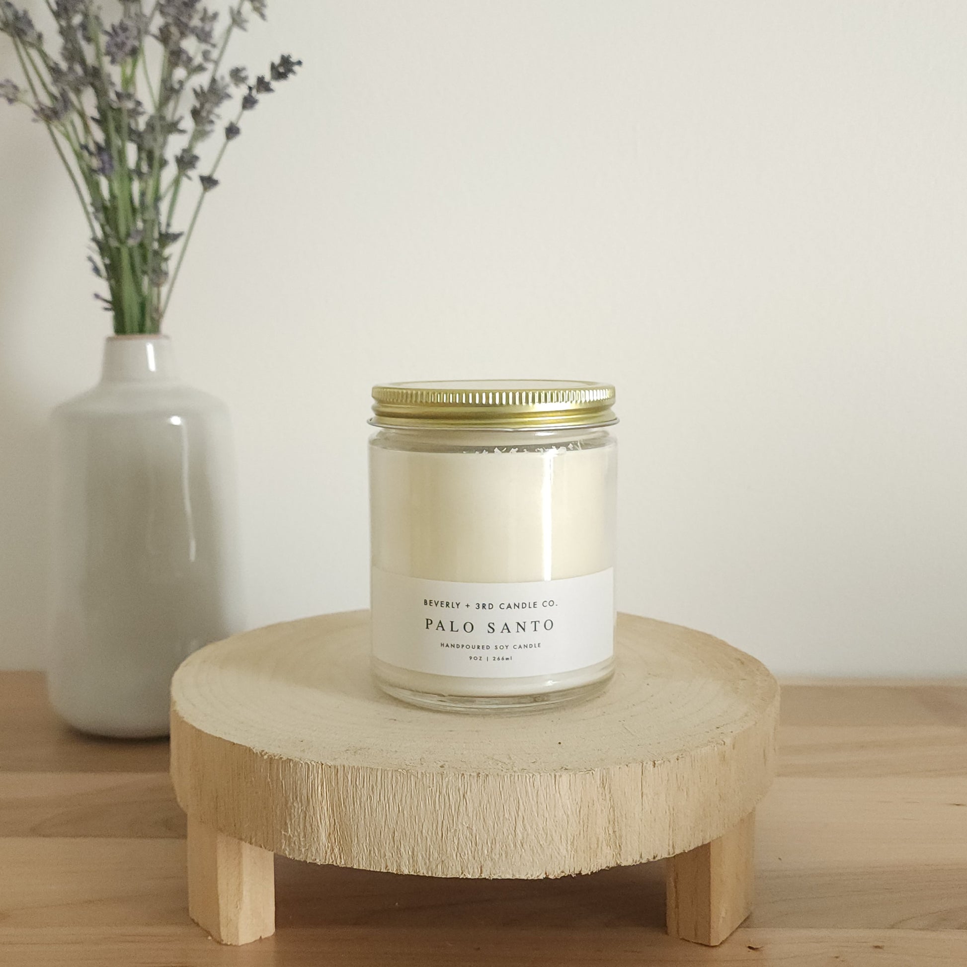 A "Palo Santo" scented soy candle in a clear glass jar with a gold lid and a white label, sits on a light wooden display stand. In the background, a white vase holds a small bouquet of lavender sprigs, against a plain white wall.