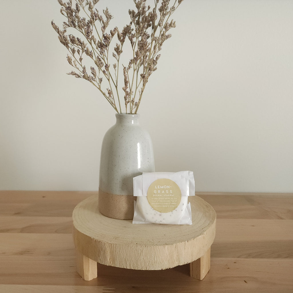 A single shower steamer in "Lemongrass" scent, individually wrapped in clear plastic with a white and yellow label, resting on a light wooden pedestal. A ceramic vase with dried botanicals is in the background.
