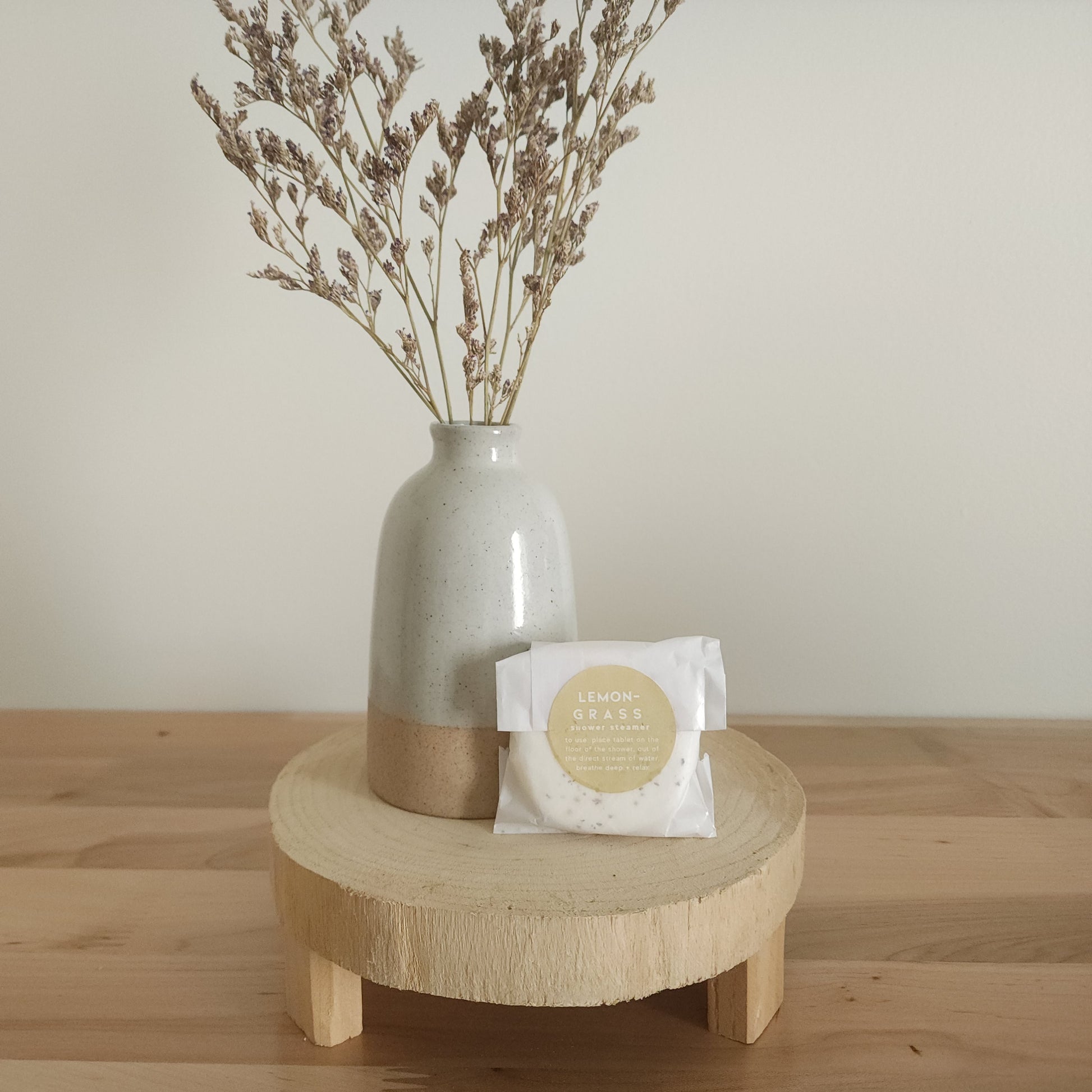 A single shower steamer in "Lemongrass" scent, individually wrapped in clear plastic with a white and yellow label, resting on a light wooden pedestal. A ceramic vase with dried botanicals is in the background.