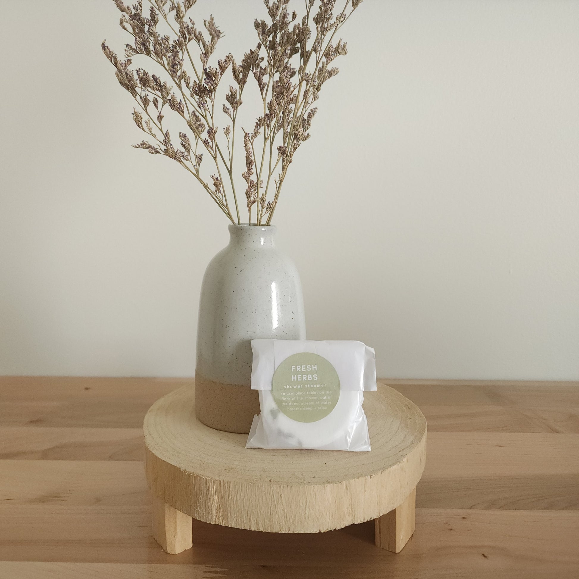 A single shower steamer in "Fresh Herbs" scent, individually wrapped in clear plastic with a white and green label, resting on a light wooden pedestal. A ceramic vase with dried botanicals is in the background.
