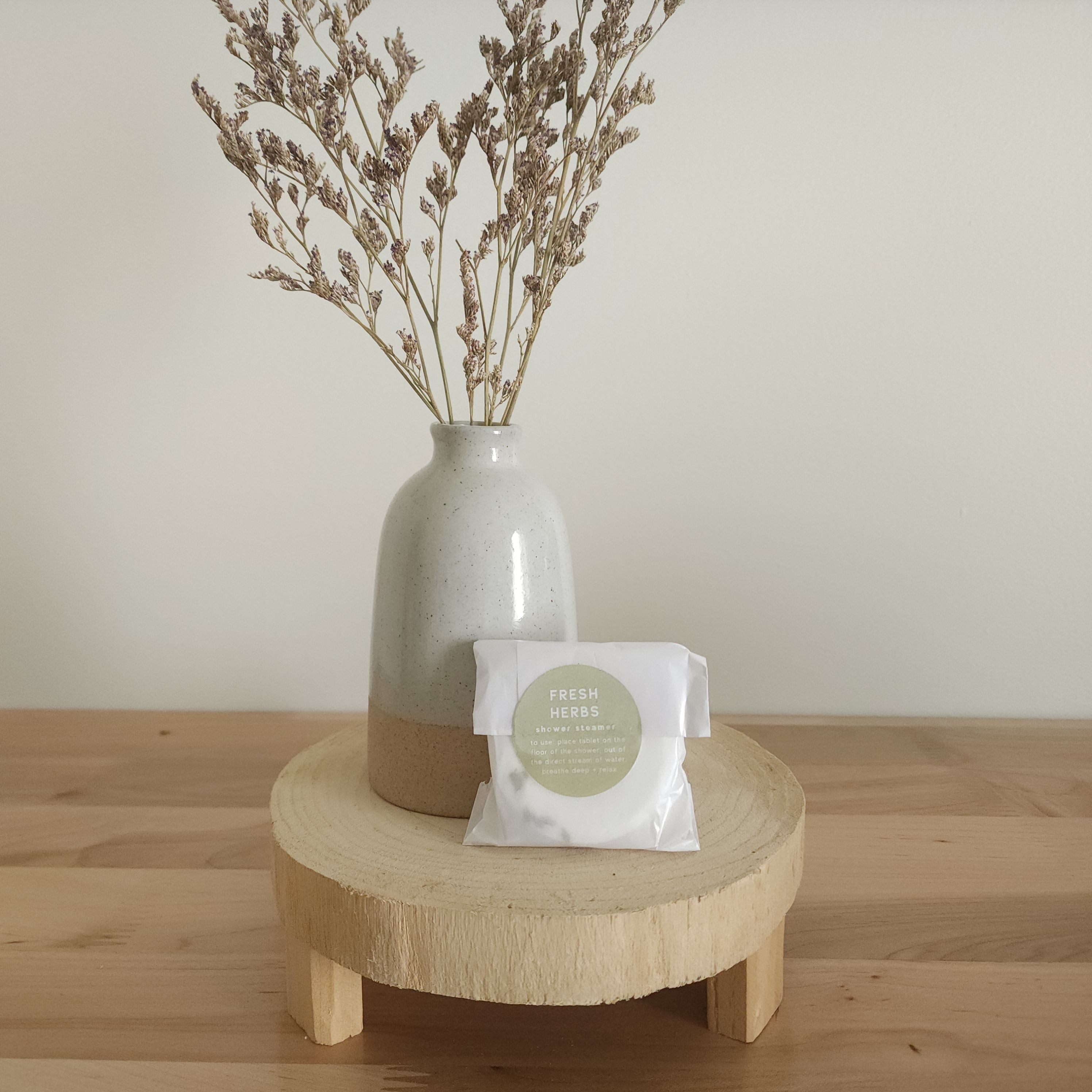 A single shower steamer in "Fresh Herbs" scent, individually wrapped in clear plastic with a white and green label, resting on a light wooden pedestal. A ceramic vase with dried botanicals is in the background.