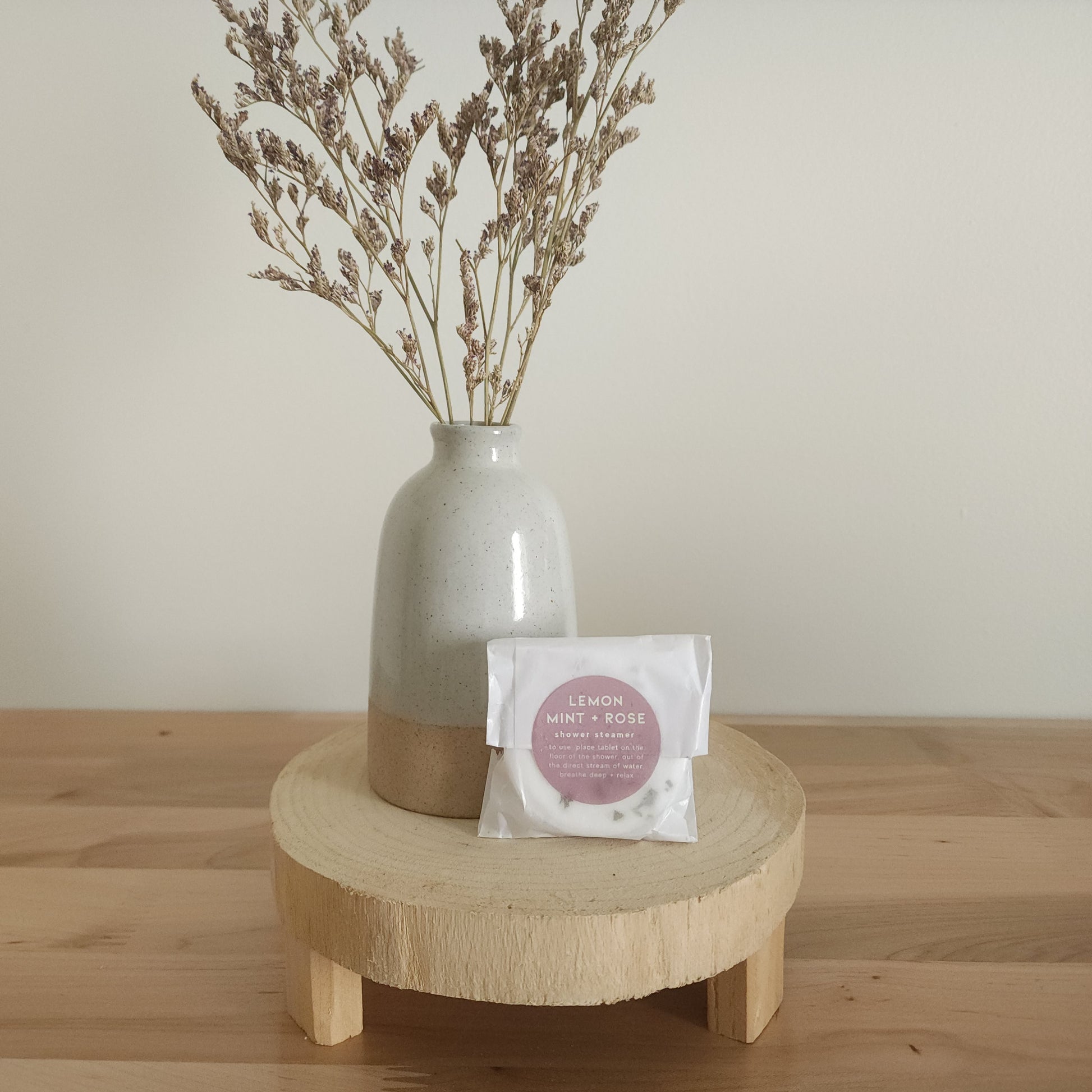 A single shower steamer in "Lemon Mint + Rose" scent, individually wrapped in clear plastic with a white and pink label, resting on a light wooden pedestal. A ceramic vase with dried botanicals is in the background.