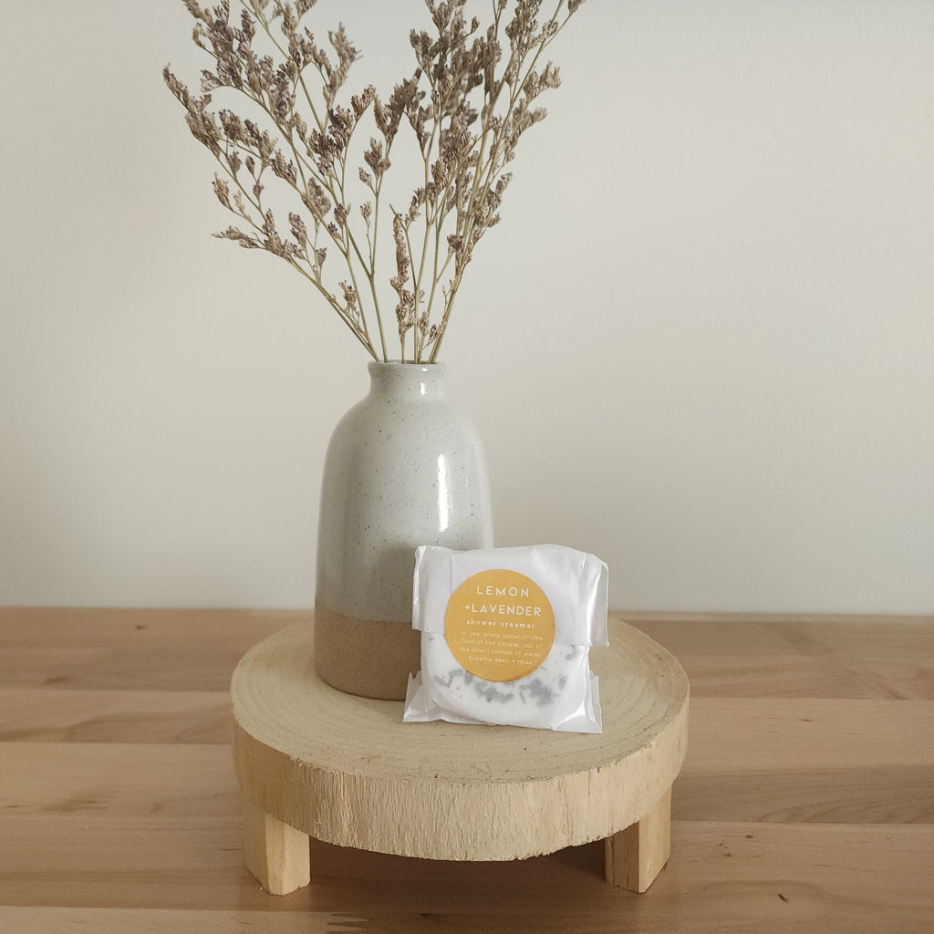 A single shower steamer in "Lemon + Lavender" scent, individually wrapped in clear plastic with a white and yellow label, resting on a light wooden pedestal. A ceramic vase with dried botanicals is in the background.