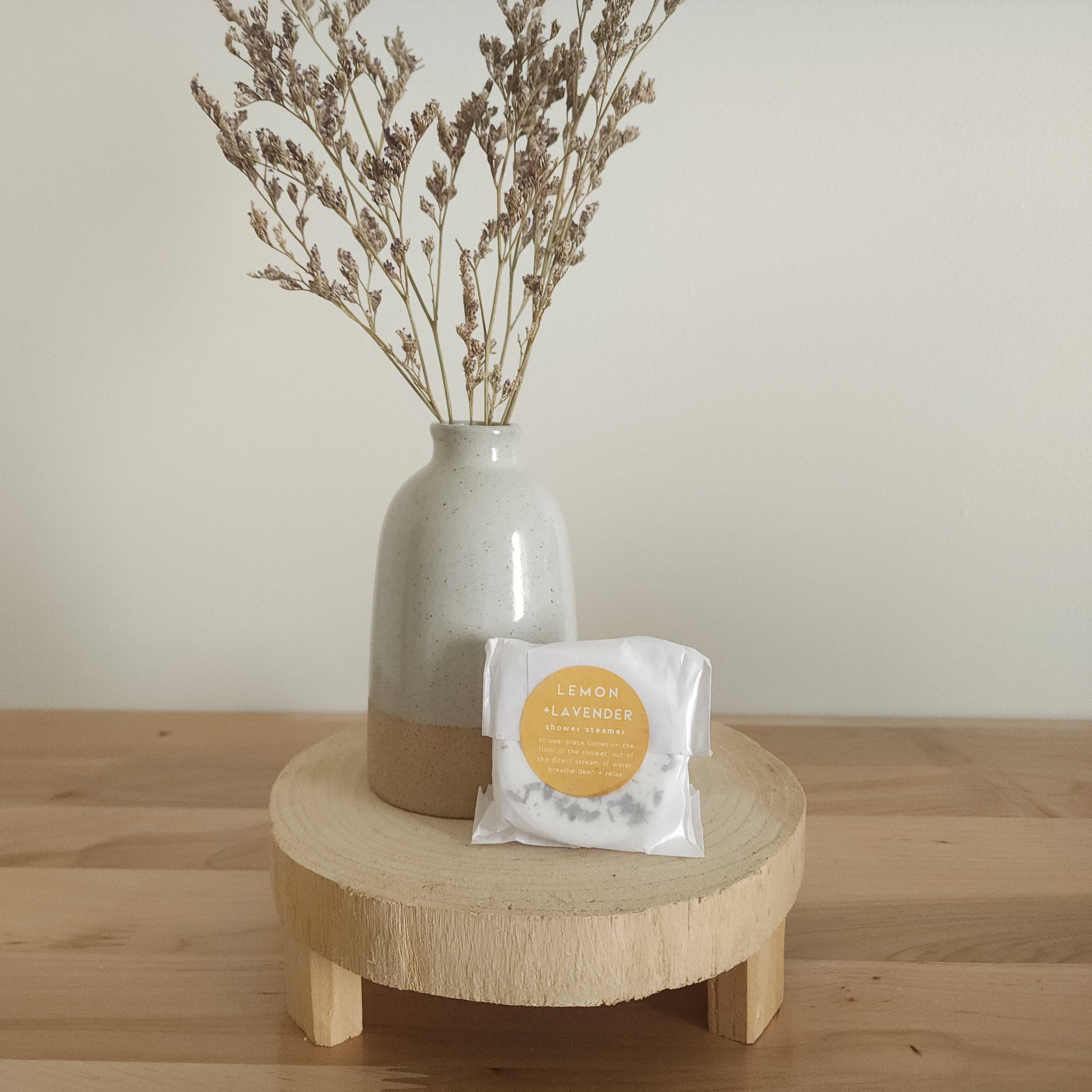 A single shower steamer in "Lemon + Lavender" scent, individually wrapped in clear plastic with a white and yellow label, resting on a light wooden pedestal. A ceramic vase with dried botanicals is in the background.
