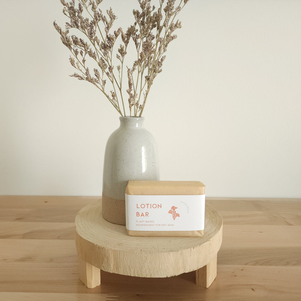  A rectangular "Lotion Bar" for dry skin, wrapped in paper with a warm peach-toned label, rests on a light wooden pedestal. A beige ceramic vase with dried botanicals is in the background, against a white wall.