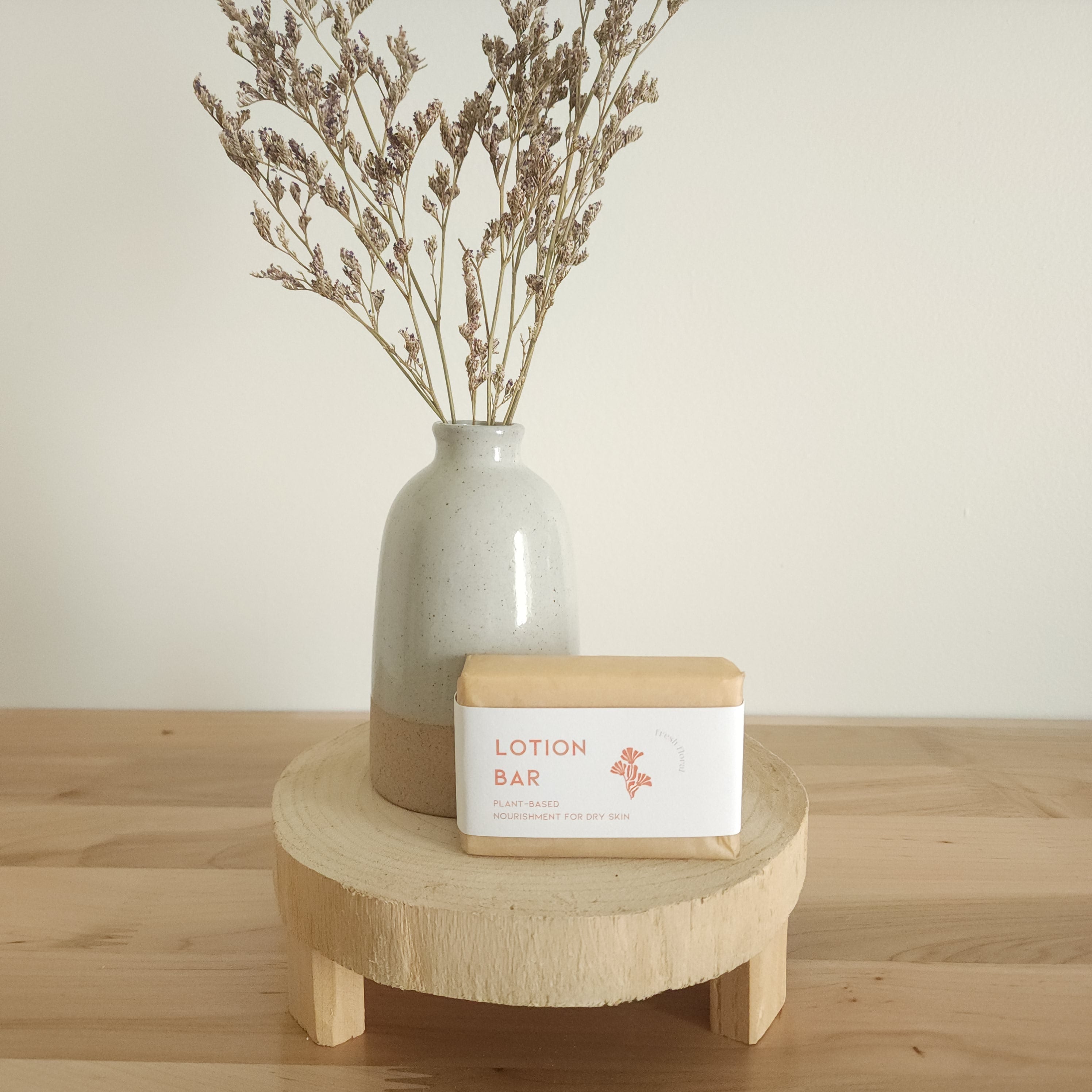  A rectangular "Lotion Bar" for dry skin, wrapped in paper with a warm peach-toned label, rests on a light wooden pedestal. A beige ceramic vase with dried botanicals is in the background, against a white wall.