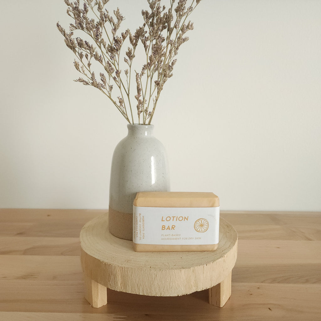 A rectangular "Lotion Bar" for dry skin, wrapped in paper with an orange citrus illustration on its label, rests on a light wooden pedestal. A beige ceramic vase with dried botanicals is in the background, against a white wall.