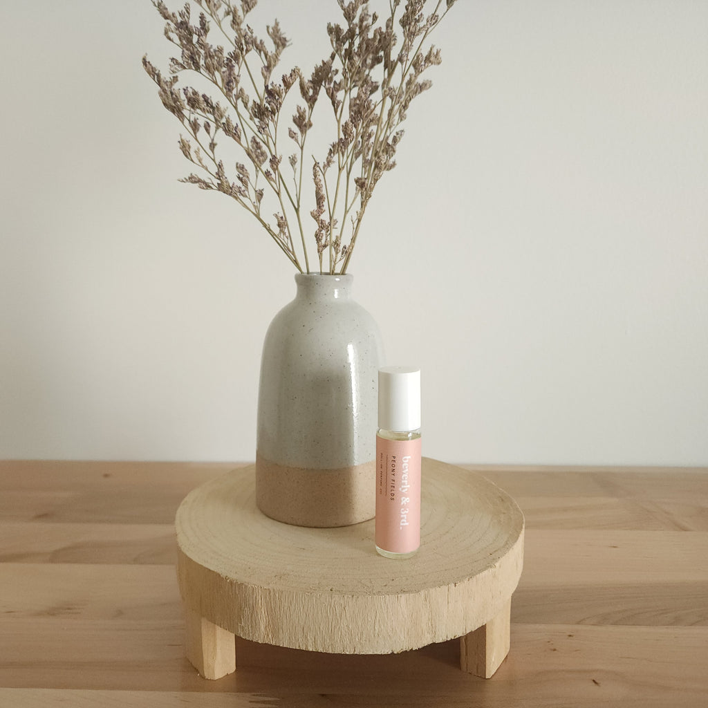 A small aromatherapy roller bottle with a "Peony Fields" label sits on a light wooden pedestal next to a beige ceramic vase holding dried botanicals, against a white background.
