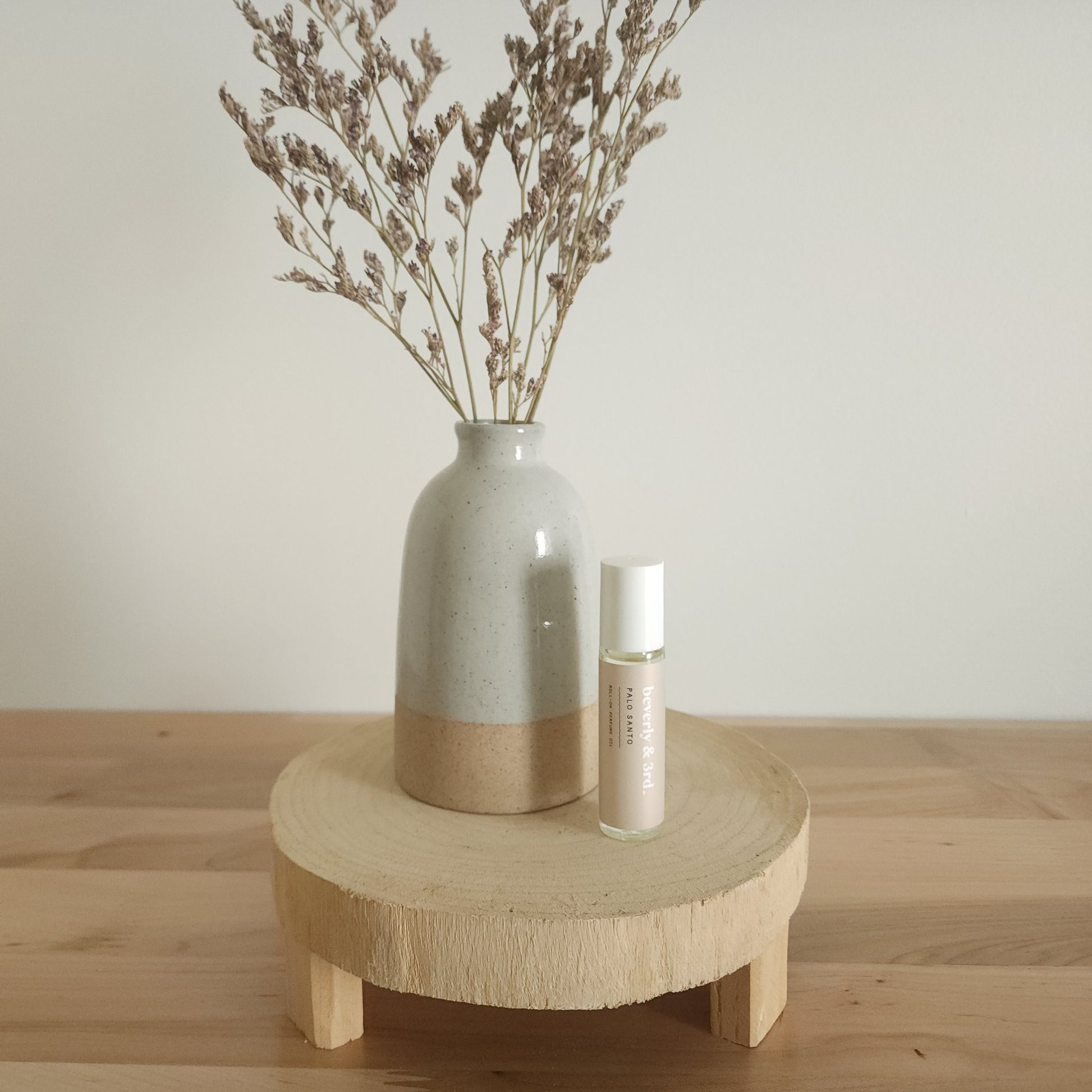 A small aromatherapy roller bottle with a "Palo Santo" label sits on a light wooden pedestal next to a beige ceramic vase holding dried botanicals, against a white background.