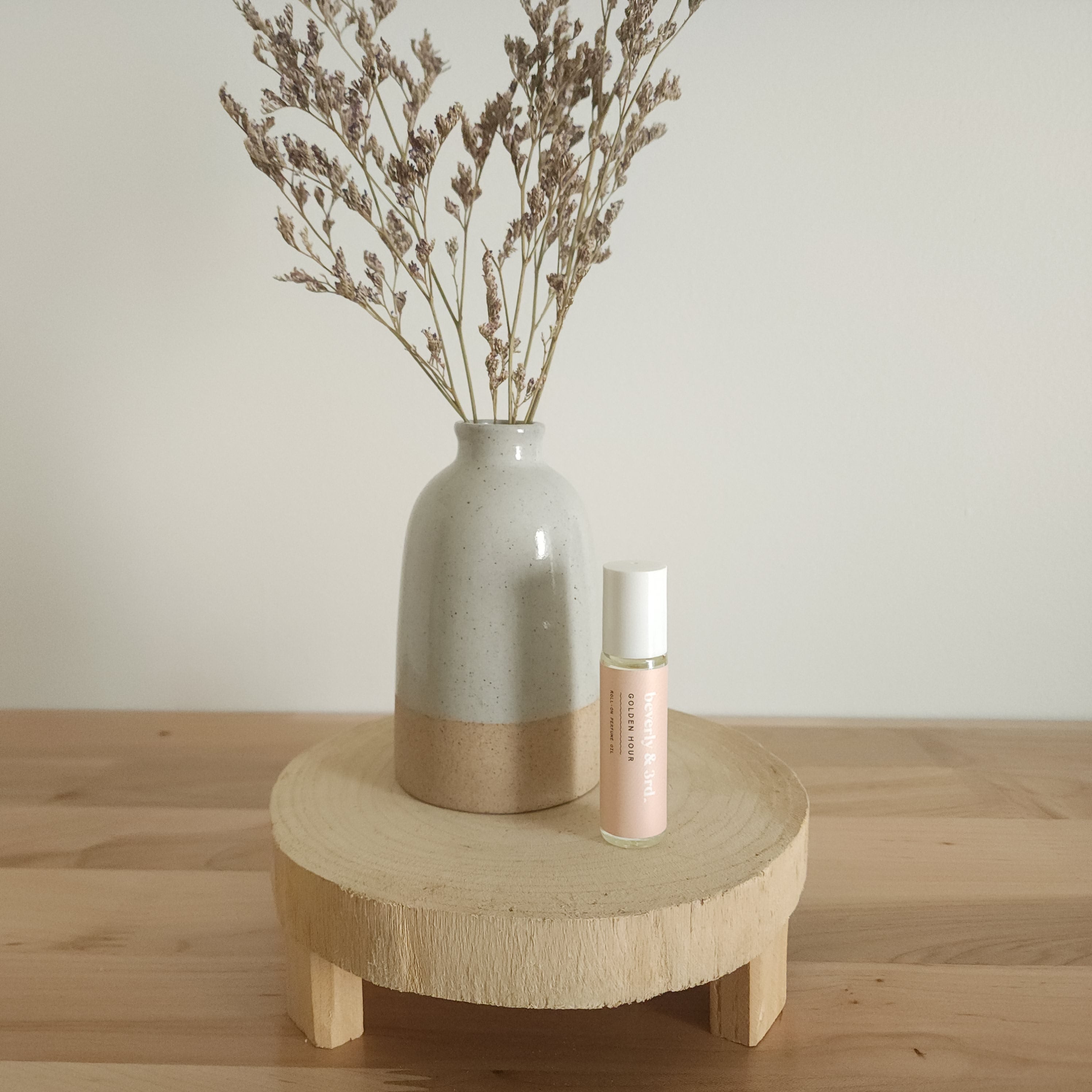 A small aromatherapy roller bottle with a "Golden Hour" label sits on a light wooden pedestal next to a beige ceramic vase holding dried botanicals, against a white background.