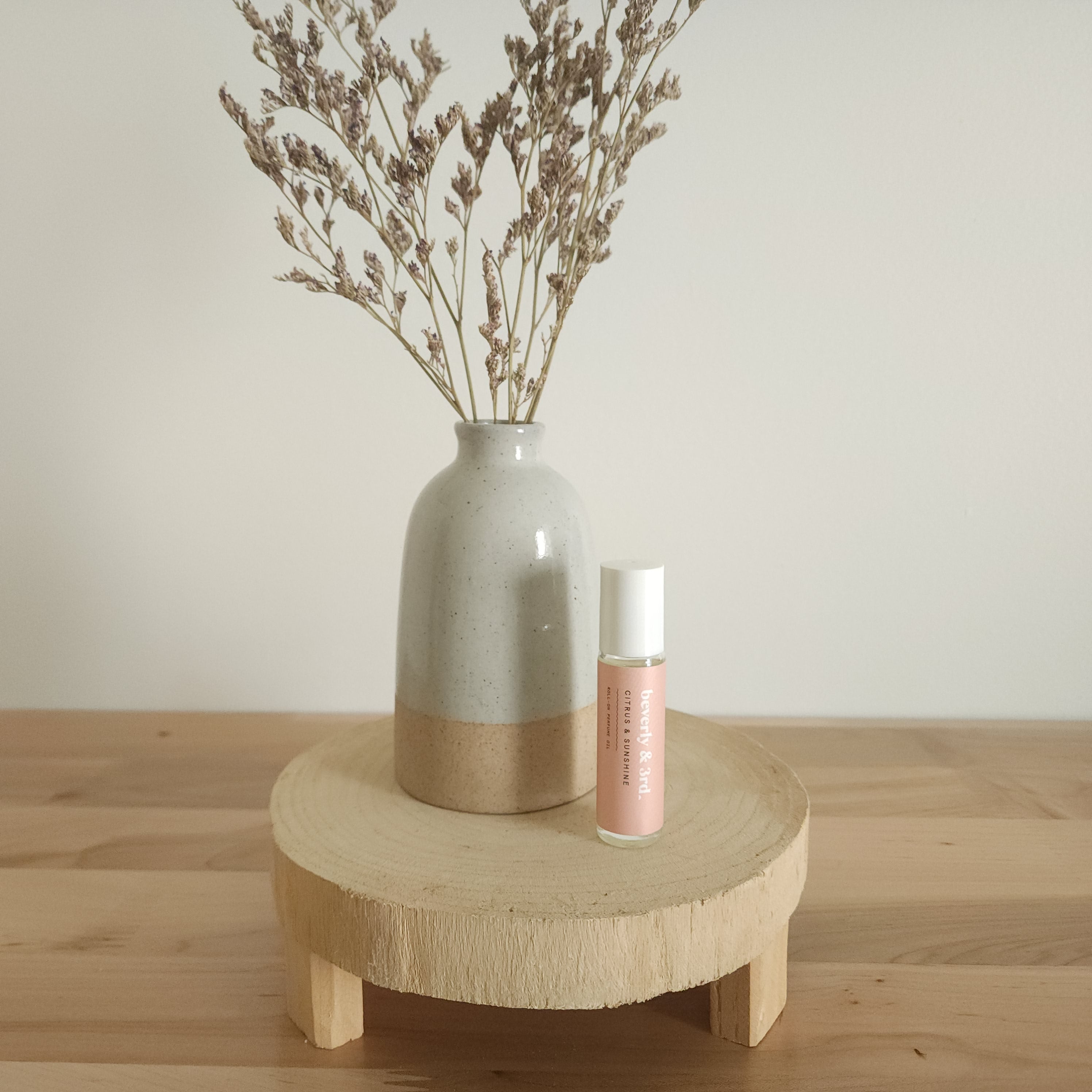 A small aromatherapy roller bottle with a "Citrus & Sunshine" label sits on a light wooden pedestal next to a beige ceramic vase holding dried botanicals, against a white background.