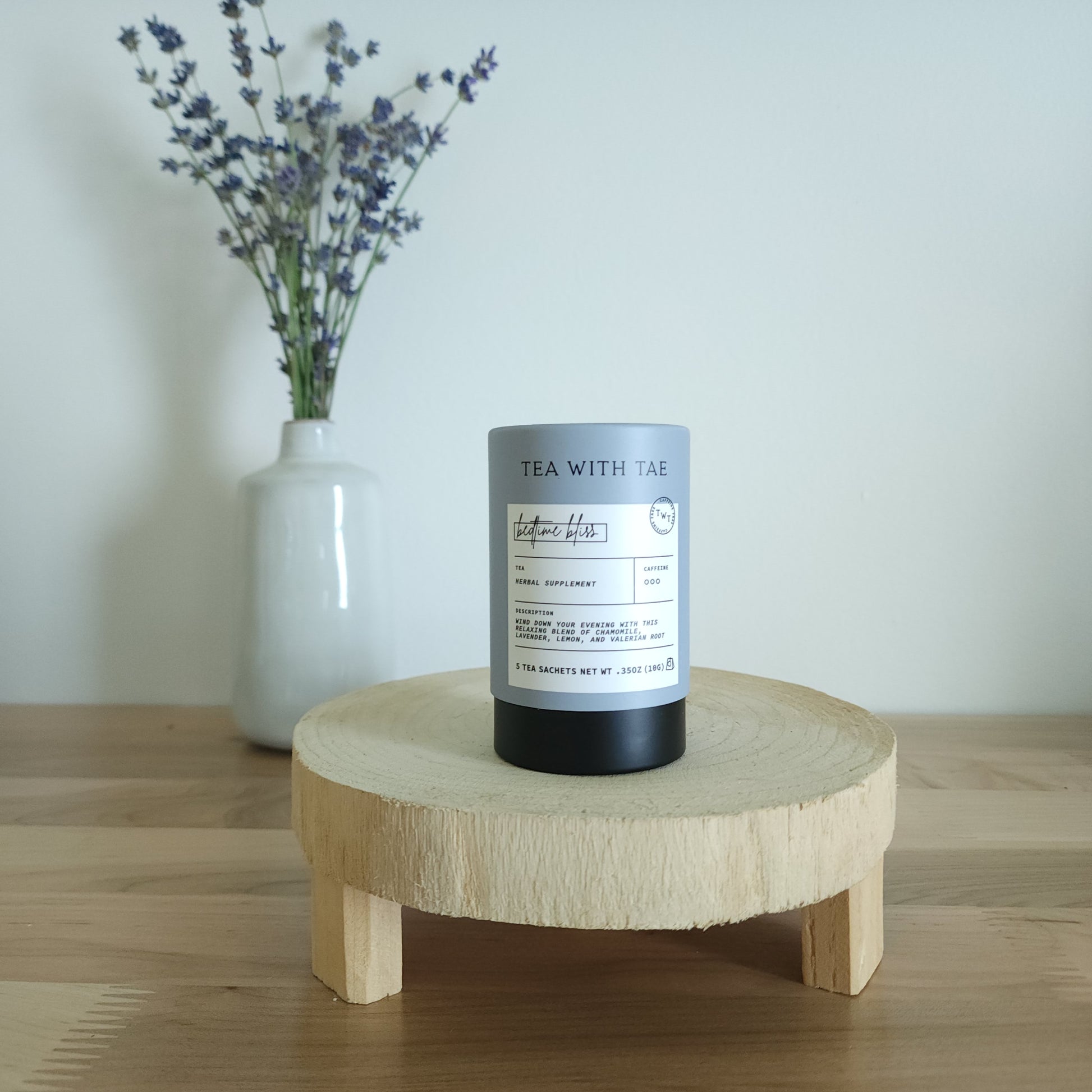 A cylindrical tea canister labeled "Tea With Tae, Bedtime Bliss Herbal Supplement" stands on a light wooden display stand. In the background, a white vase holds a small bouquet of lavender sprigs, against a plain white wall.
