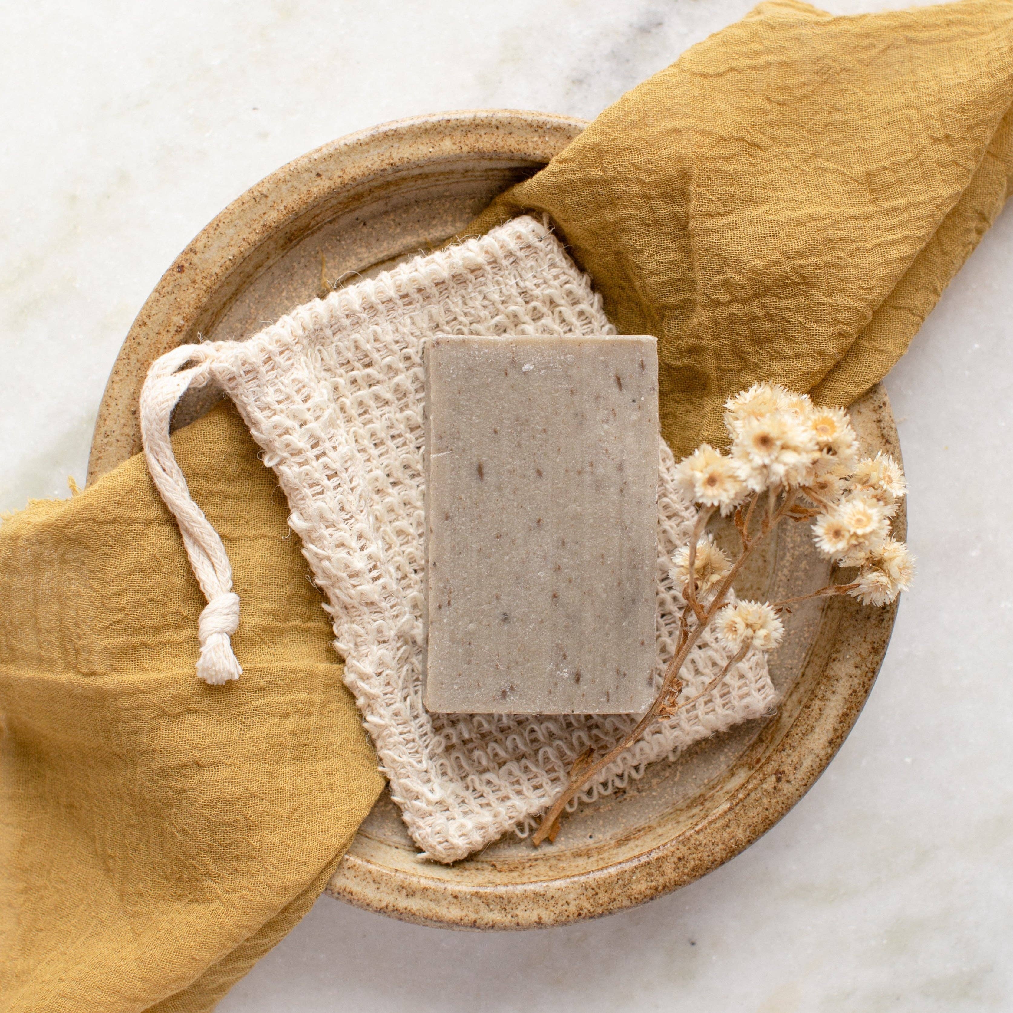 A rectangular bar of natural grey soap rests on an exfoliating soap saver bag, all placed on a textured ceramic plate. A mustard yellow linen cloth and a sprig of dried white flowers complete the rustic, natural bathroom aesthetic.