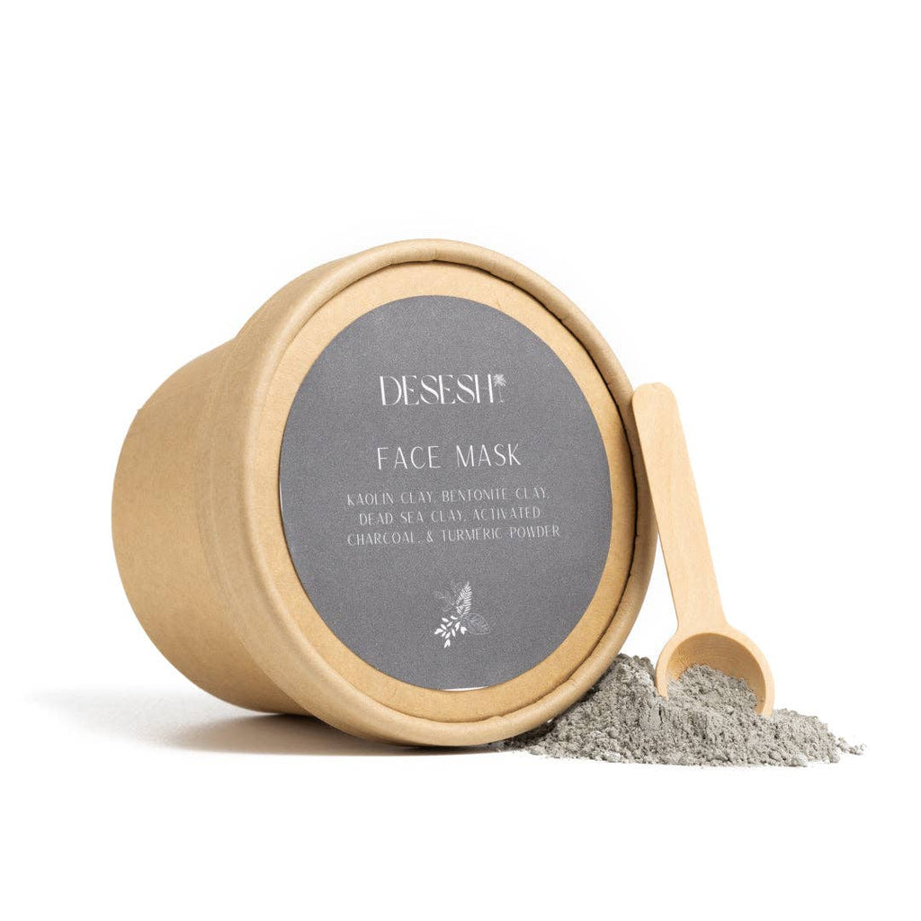 Round kraft paper container labeled 'DESESH FACE MASK' with ingredients listed, including kaolin clay, bentonite clay, dead sea clay, activated charcoal, and turmeric powder, next to a small wooden scoop and a pile of gray powder on a white background.