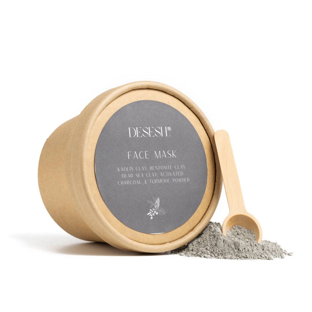 Round kraft paper container labeled 'DESESH FACE MASK' with ingredients listed, including kaolin clay, bentonite clay, dead sea clay, activated charcoal, and turmeric powder, next to a small wooden scoop and a pile of gray powder on a white background.