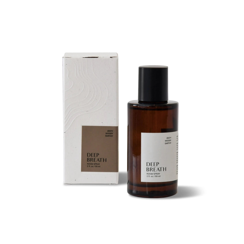 Brown glass bottle of Deep Breath Room Spray by The Glow Co, with black spray nozzle and cap off to the side, sitting on a textured gray surface. The bottle's label reads 'Deep Breath' and 'Zesty Woody Earthy'. A small white dish holding a clear crystal and a dark stone is also visible. Soft, white, rounded furniture is blurred in the background.