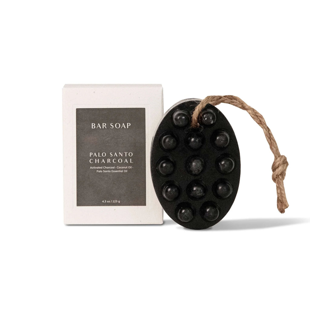 Oval black bar soap with raised bumps and a looped brown string, labeled 'Palo Santo Charcoal' and 'Activated Charcoal Coconut Oil Palo Santo Essential Oil', next to a white box labeled 'BAR SOAP' and 'PALO SANTO CHARCOAL'. Both items are on a white background.
