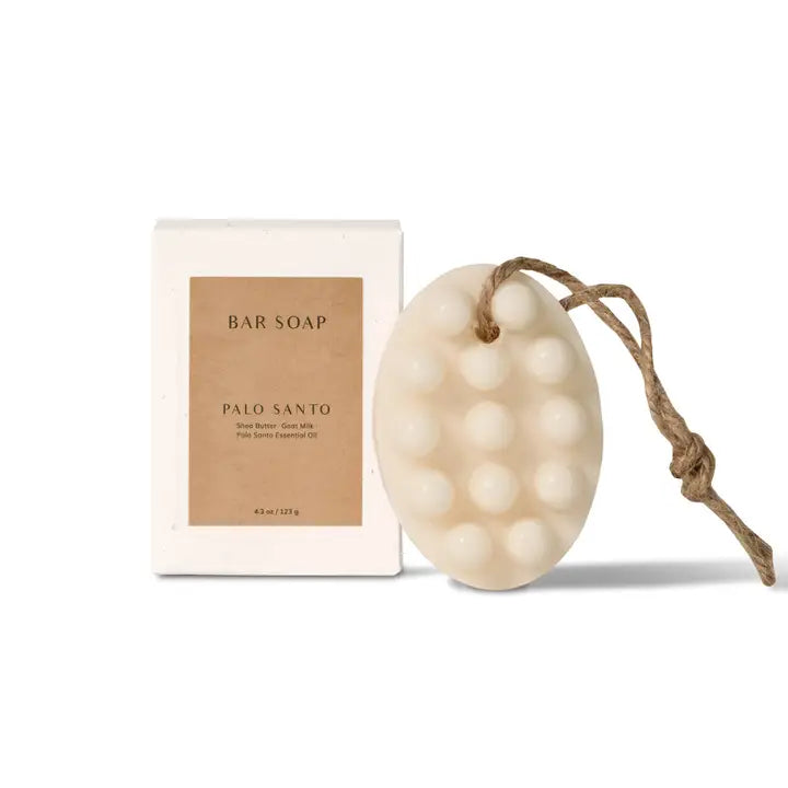 Oval cream-colored bar soap with raised bumps and a looped brown string, labeled 'Palo Santo' and 'Shea Butter - Goat Milk - Palo Santo Essential Oil', next to a white box labeled 'BAR SOAP' and 'PALO SANTO'. Both items are on a white background.