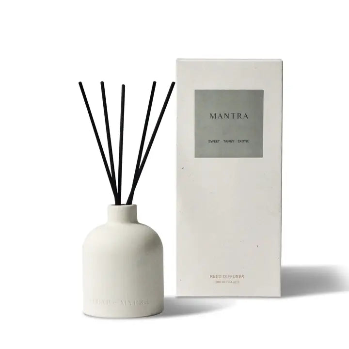 White ceramic diffuser bottle with black reeds, labeled 'MANTRA' and 'SWEET TANGY EXOTIC', next to a white box labeled 'MANTRA' and 'REED DIFFUSER'.