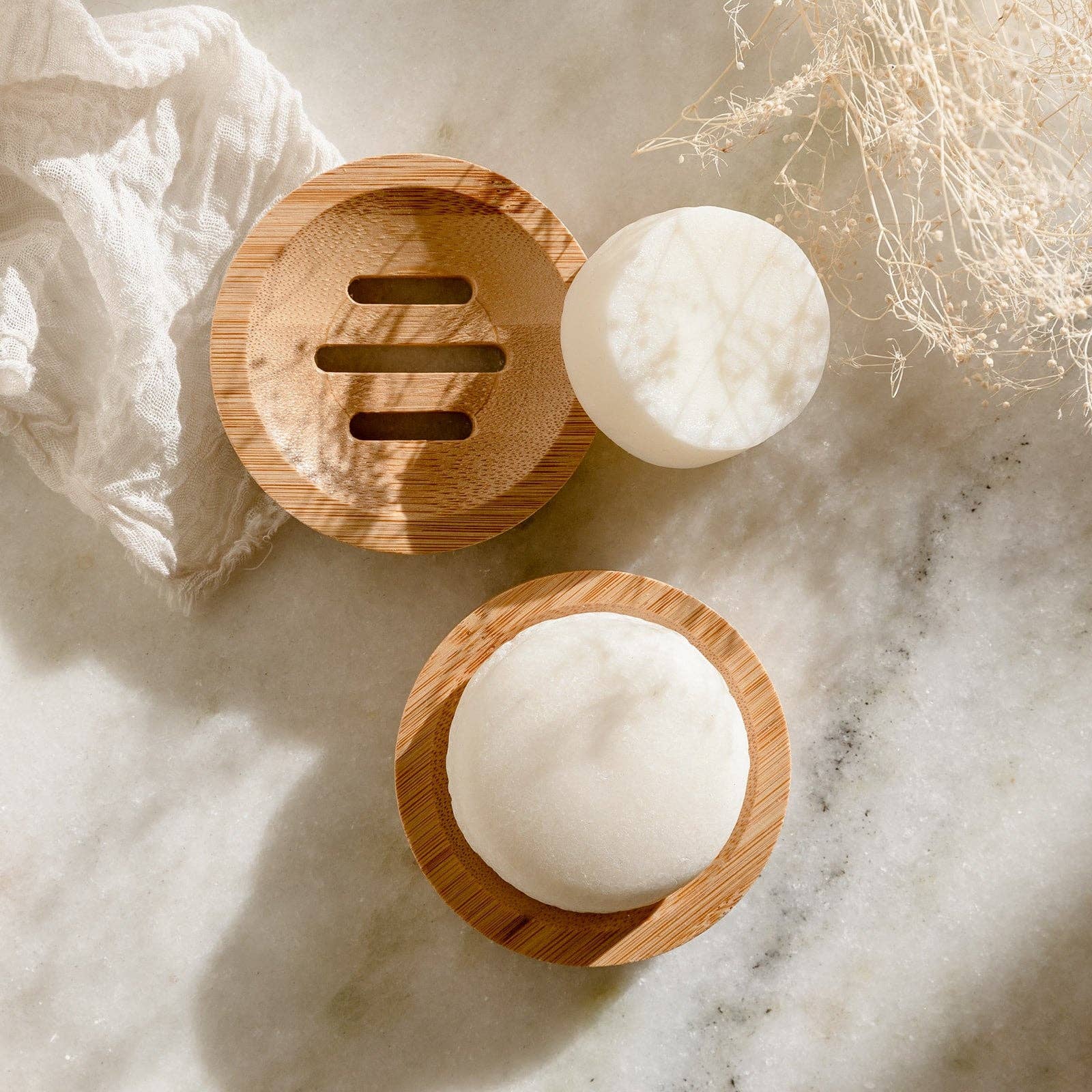 Two round, white shampoo or conditioner bars sit on separate bamboo soap dishes. One dish is empty, while the other holds a bar with its top visible. The items are arranged on a light marble surface with dappled sunlight and a piece of white textured fabric, creating a serene, natural aesthetic.